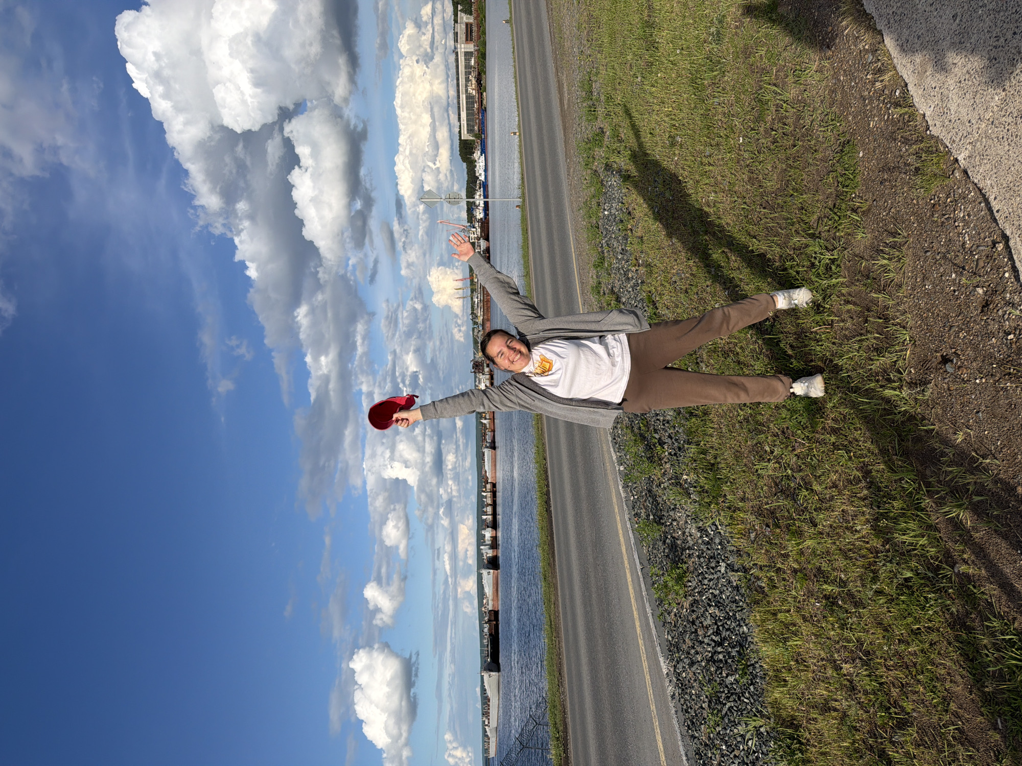 A person lies on their back beside a two-lane road in Chaikovsky, Russia. The person is wearing brown pants, a white t-shirt, a gray sweater, and white sneakers. Their arms are outstretched, holding a red baseball cap in their left hand. The road runs parallel to a body of water with industrial buildings on the opposite shore. A grassy shoulder lies between the road and a concrete barrier. The sky is blue with puffy white clouds.
