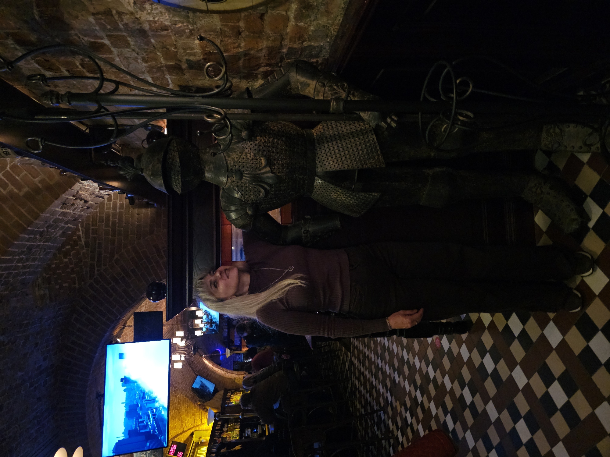 A medium shot captures a blonde woman in a purple turtleneck sweater and dark pants standing next to a full-body suit of armor in a dimly lit establishment with a brick arched ceiling. The woman gazes upwards and to her right, her left hand resting on the armor. The floor features a checkered pattern of black, white, brown, and beige tiles. In the background, a large screen displays a cityscape, and shelves with bottles are visible behind a bar. The overall ambiance suggests a bar or restaurant with a medieval or historical theme. The location is identified as Moscow, Russia.