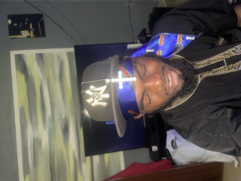 A man is lying down, dressed in a black robe with gold trim. He wears a black baseball cap with a logo on it, a blue velvet headband with green and red stripes, and a cross made of small white stones. He has a full beard and is smiling. Behind him, there's a television, a gray painting with yellow and white streaks, and a smaller black and white picture. A white sheet and some red fabric are also visible.