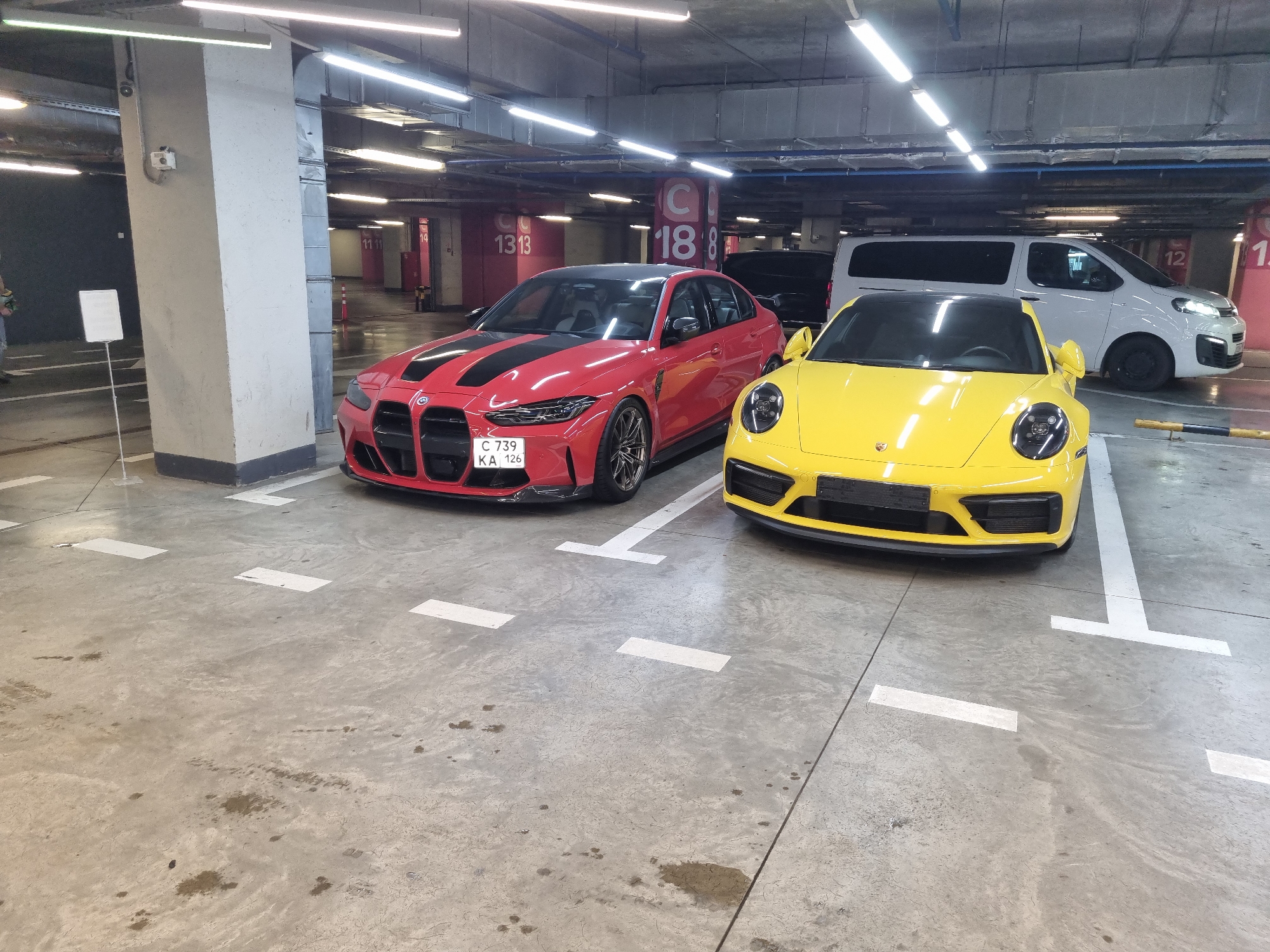 This image depicts an indoor scene within an underground or multi-story parking garage, likely located in Sochi, Russia. Two high-performance sports cars are prominently featured in the foreground, parked side-by-side.
On the left, a red BMW M3 or M4 (G80/G82 generation) is parked, distinguished by its large black kidney grilles, black racing stripes adorning its hood, and a black roof. It is equipped with black multi-spoke alloy wheels and red brake calipers. The Russian license plate, "C 739 KA 126", is clearly visible on its front bumper.
To its right, a bright yellow Porsche 911 (likely a 992 generation) is parked, facing the viewer. This car also features black accents on its front bumper and multi-spoke black wheels.
Behind the yellow Porsche, a white commercial or passenger van, possibly a Citroën Jumpy, is visible. Other vehicles are partially seen in the background, including a dark-colored car behind the red BMW.
The environment consists of a concrete floor with white parking space markings and some dark stains. The ceiling is illuminated by numerous fluorescent strip lights, and ventilation ducts are apparent. Large concrete pillars, marked with numbers such as "C 18 8", "13 13", "11 11", "14", and "12", indicate parking sections. To the far left, a white, blank sign on a stand is visible, along with the lower body of a person. The scene portrays a well-lit, functional parking facility housing luxury automobiles.