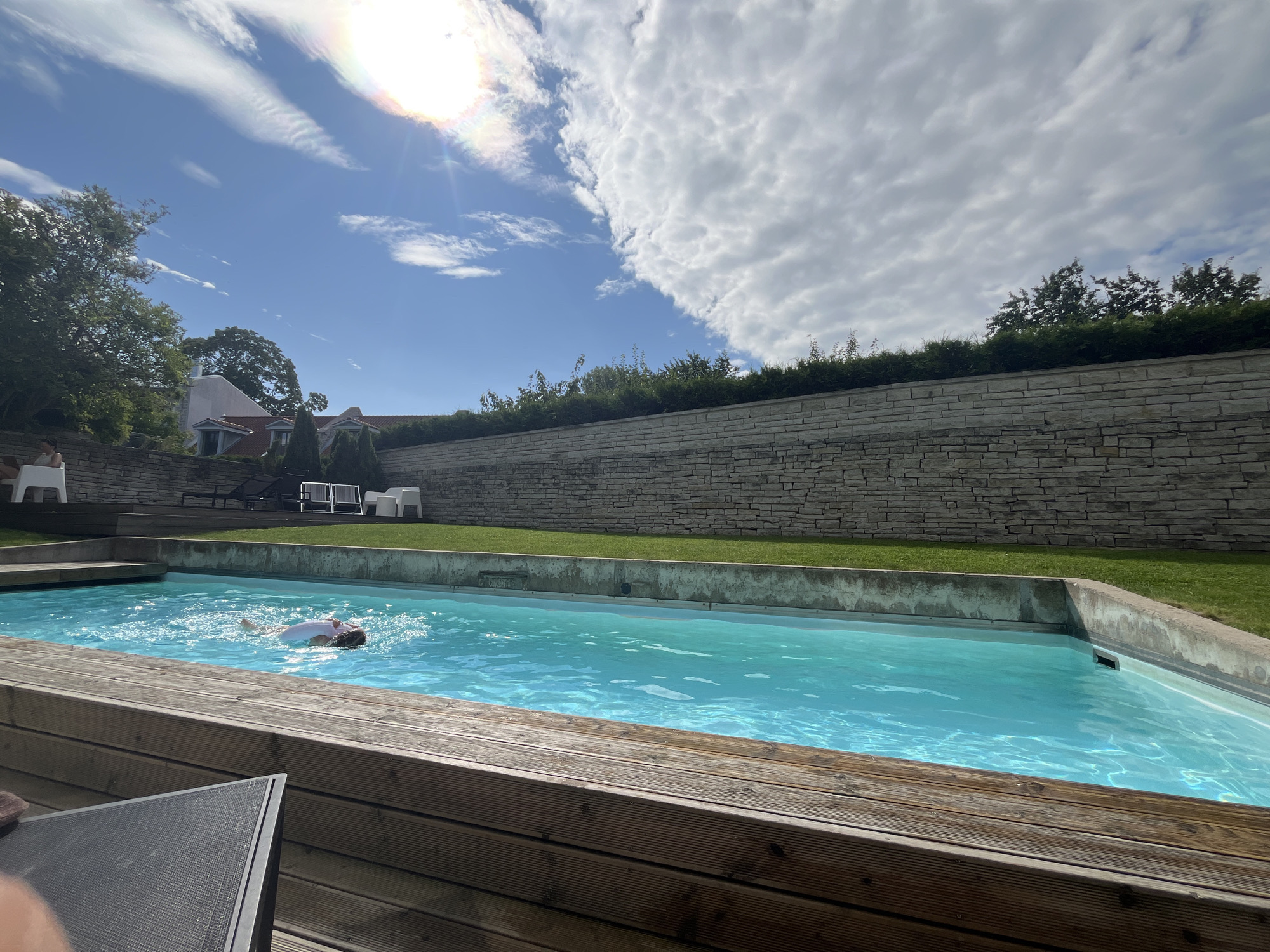A bright, sunny day is captured in this image showing an outdoor swimming pool located in Kuressaare, Estonia. The pool is filled with clear turquoise water and is surrounded by a wooden deck in the foreground. In the middle of the pool, a person is swimming, appearing to be doing the backstroke with their arms extended. The pool area is enclosed by a low stone wall, behind which a manicured lawn and trees are visible. In the background, houses with red-tiled roofs can be seen, nestled amongst the trees. The sky is a vibrant blue with scattered white clouds, and the sun is shining brightly, casting reflections on the water's surface. A sun halo is visible in the upper left corner of the image.