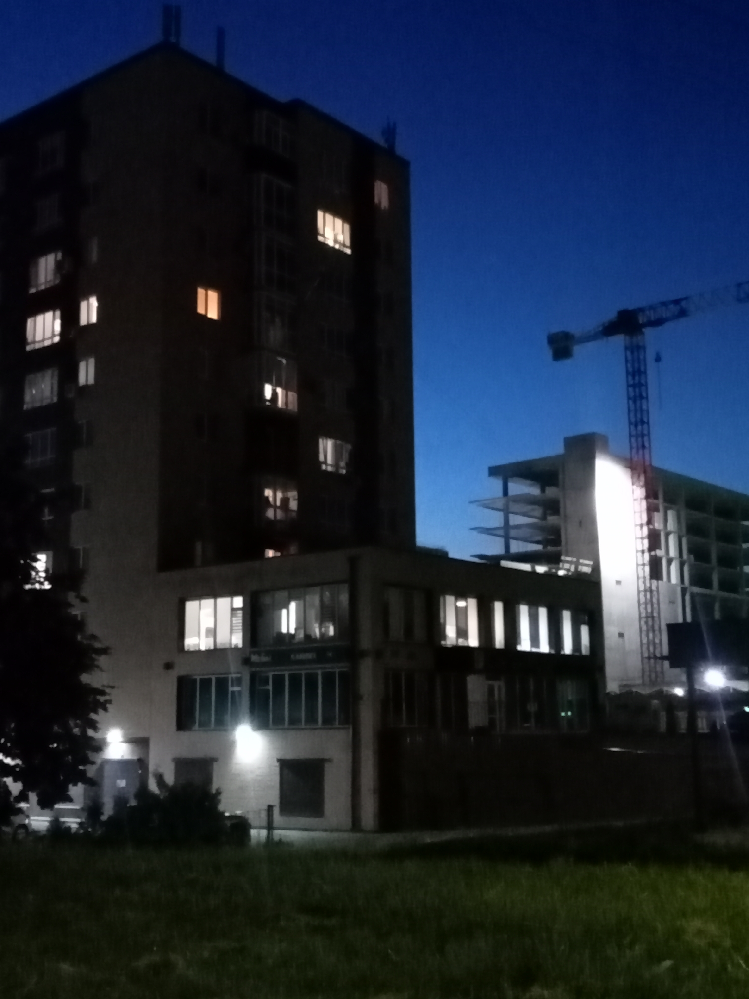 In the city of Unknown, Unknown, a twilight scene unfolds. A tall, dark residential building stands prominently on the left, its windows illuminated with warm, artificial light, suggesting occupancy. To the right, a construction site is visible, with a towering crane silhouetted against the deepening blue sky, and a partially constructed building revealing its skeletal frame. The ground in the foreground is covered with grass, casting long shadows in the low light. The overall impression is one of urban transition, with residential life coexisting alongside ongoing development.