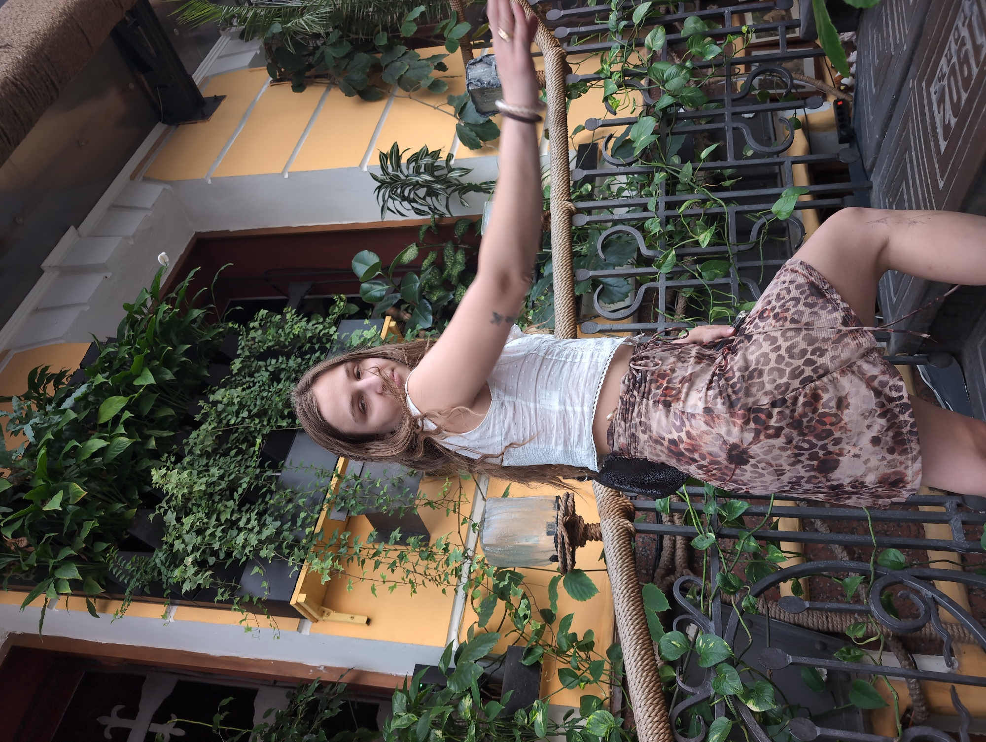 A young woman with long brown hair and fair skin is posing outdoors in front of a yellow building covered in lush green plants. She is wearing a white cropped top with an intricate pattern and a tan and brown leopard-print skirt. Her left arm is extended to the right, with her hand slightly blurred as if in motion. She is looking towards the camera with a slight smile. The background features a railing, a rope, and more plants, suggesting an outdoor cafe or a restaurant setting. The image does not provide enough information to determine the specific location within Kyiv, Ukraine.