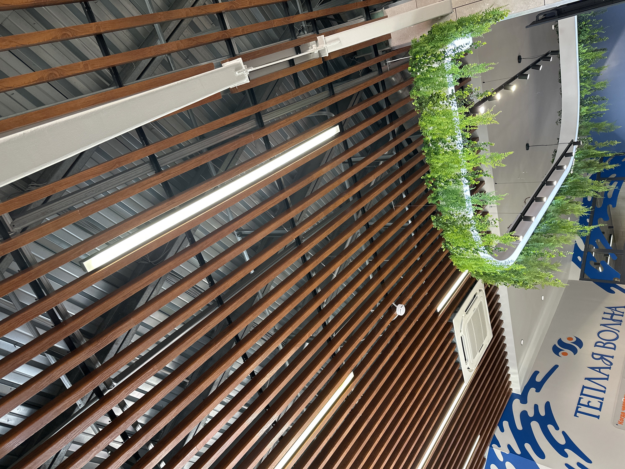 A low-angle shot showcases the architectural details of what appears to be an interior space, likely within a commercial building or shopping center in Votkinsk, Russia. The ceiling is composed of parallel wooden slats, giving a sense of depth and direction to the image. Interspersed among the slats are light fixtures, providing illumination to the area.
To the right, a structure or signboard can be seen, adorned with greenery. This structure also features a logo with the words "ТЕПЛАЯ ВОЛНА" (Warm Wave) in Russian, along with blue wave-like patterns. The overall impression is that the design incorporates natural and aquatic themes.