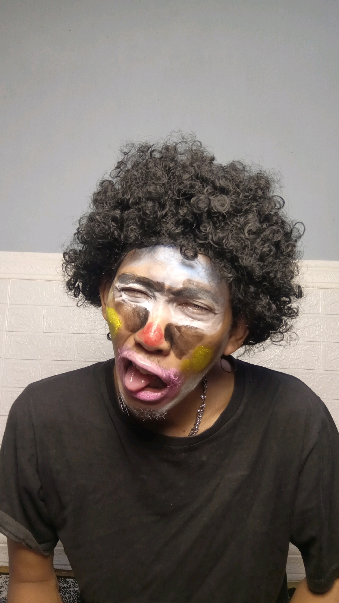 A person with a curly black wig and clown makeup is sticking their tongue out and squinting their eyes. The makeup includes white face paint, a red nose, brown patches around the eyes and cheeks, and yellow glitter on the cheeks. The person is wearing a black t-shirt and a silver chain necklace. The background appears to be a wall with white textured panels. The image does not provide any information about the location in Kebumen, Indonesia.