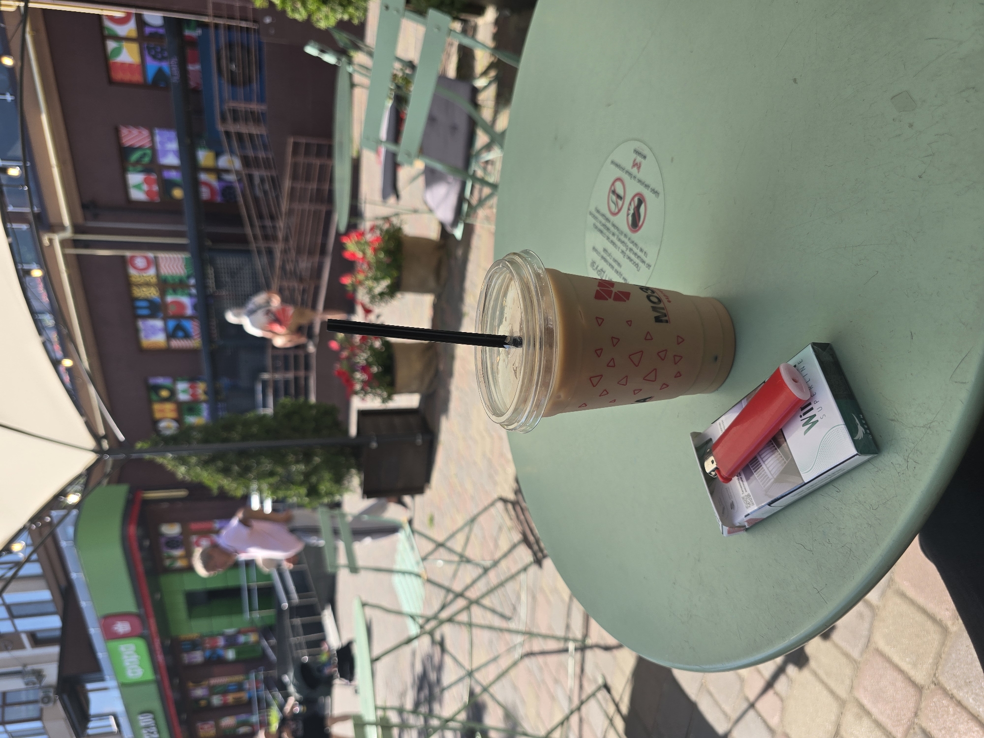 An outdoor cafe scene in Petropavlivska Borshchahivka, Ukraine features a light green, round table in the foreground. On the table rests a clear plastic cup with a beige beverage and a black straw, decorated with red triangles. Next to the cup is a pack of white and green "Winston Superline" cigarettes, topped with a red disposable lighter. In the background, people are visible walking near buildings with green and brown facades. One building displays colorful square artwork. Several light green metal chairs and tables are scattered throughout the area, and potted plants with red flowers add a touch of color. The ground is paved with interlocking bricks.