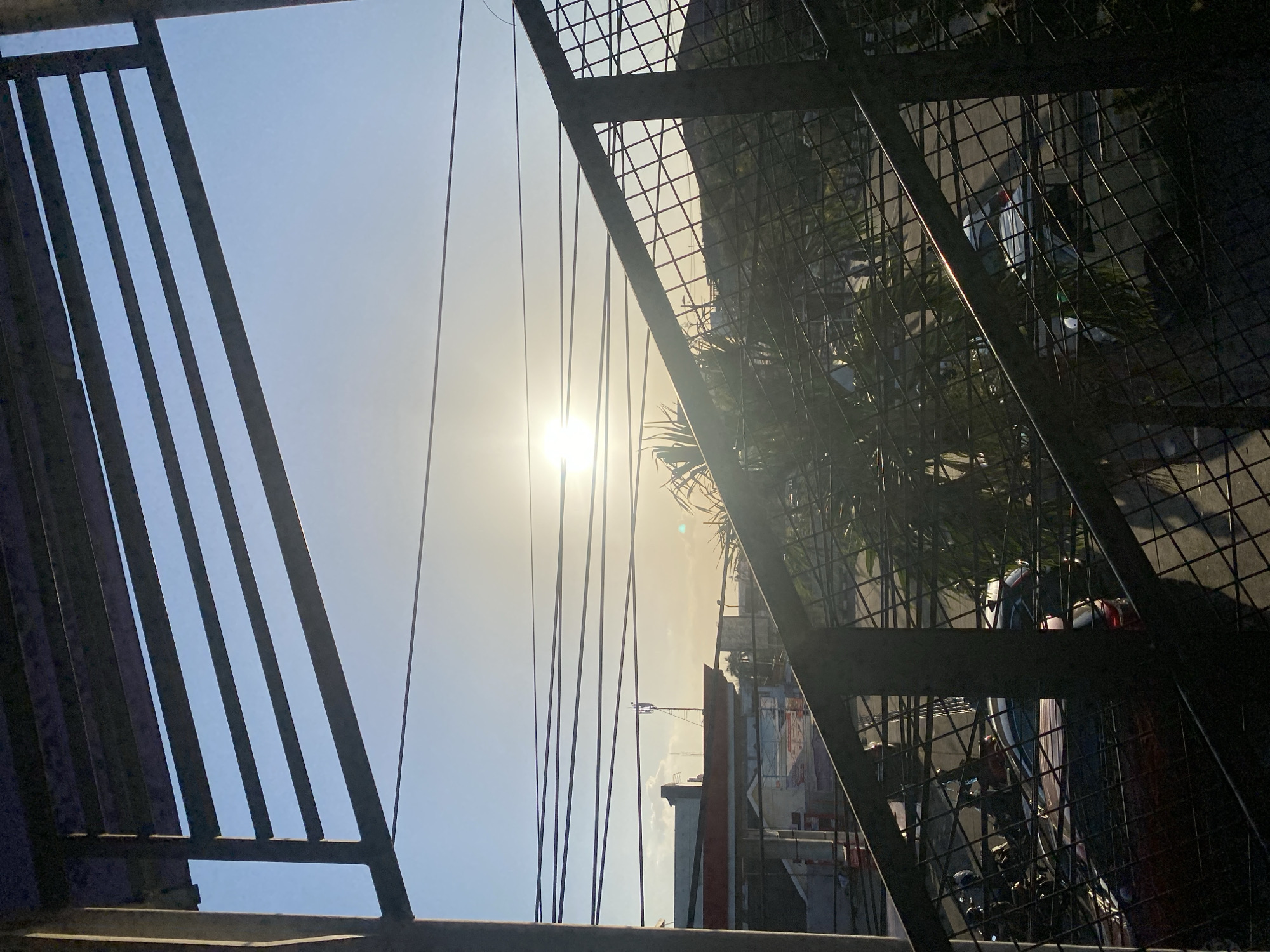 The image depicts a street view in Banjarbaru, Indonesia, during a sunset. The foreground is dominated by a metal staircase railing and overhead bars, creating a layered perspective. The sky is a clear, pale blue transitioning to a warm yellow near the horizon where the sun is setting, casting a bright glow. Several power lines crisscross the sky. In the middle ground, buildings with red roofs are visible, along with a tall communication tower. The lower portion of the image shows parked cars and motorcycles, partially obscured by the shadows and the fencing. Lush green foliage, likely palm trees, adds a tropical element to the scene. The overall atmosphere is that of an urban environment bathed in the soft light of dusk.