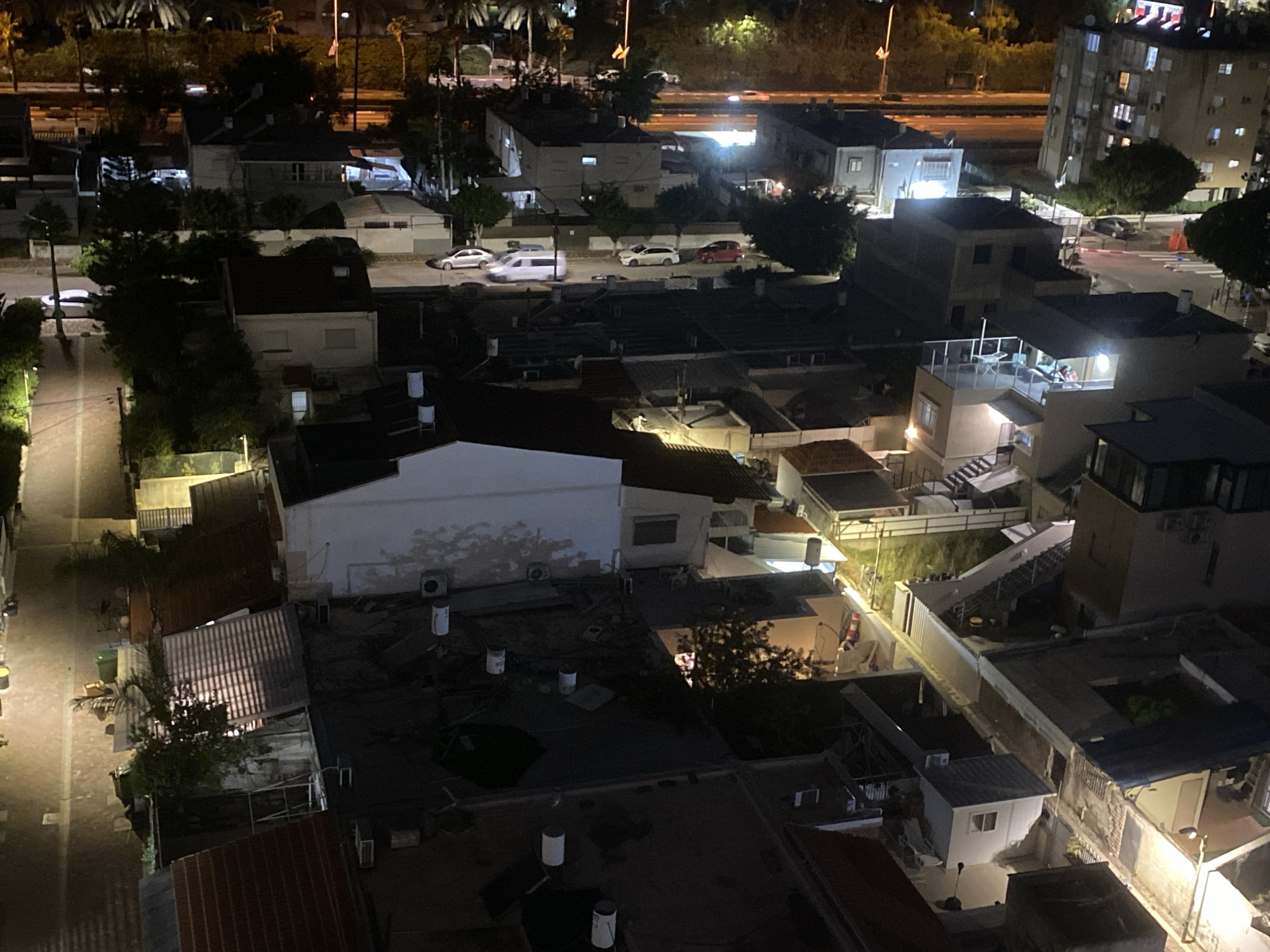 An aerial view at night in Kiryat Bialik, Israel, shows a dense residential area. Buildings vary in height, with a mix of low-rise structures and a taller apartment building. Streets are illuminated by streetlights and vehicle headlights, with a few vehicles visible. Rooftops are cluttered with objects like water tanks and air conditioning units. A light fixture on the right side is on. People are visible on a rooftop patio.