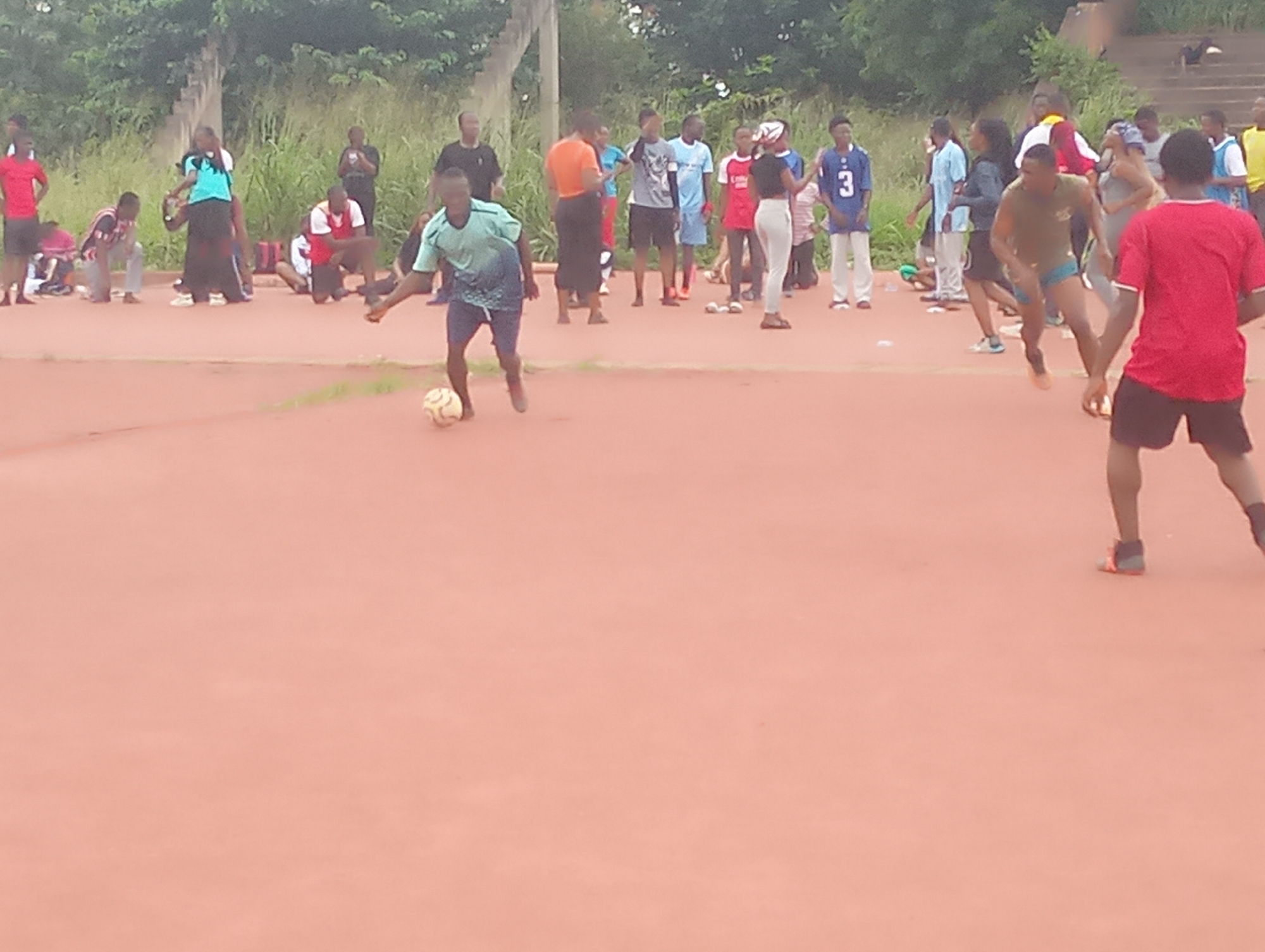 The image depicts an outdoor scene at the Nsukka city stadium in Nigeria. Several individuals, primarily young adults, are gathered on a reddish-orange athletic track. In the foreground, a young man in a blue and green t-shirt and dark shorts is actively playing soccer, dribbling a ball with his left foot. His body is in motion, suggesting he is in the process of kicking the ball. Behind him, a group of people are observing the game, some of whom are standing and others are crouching. To the right, a young man in a red t-shirt and dark shorts is also engaged in the activity, facing away from the viewer. The background shows lush green vegetation and a concrete structure, possibly a bleacher or wall, characteristic of a stadium environment. The weather appears overcast.