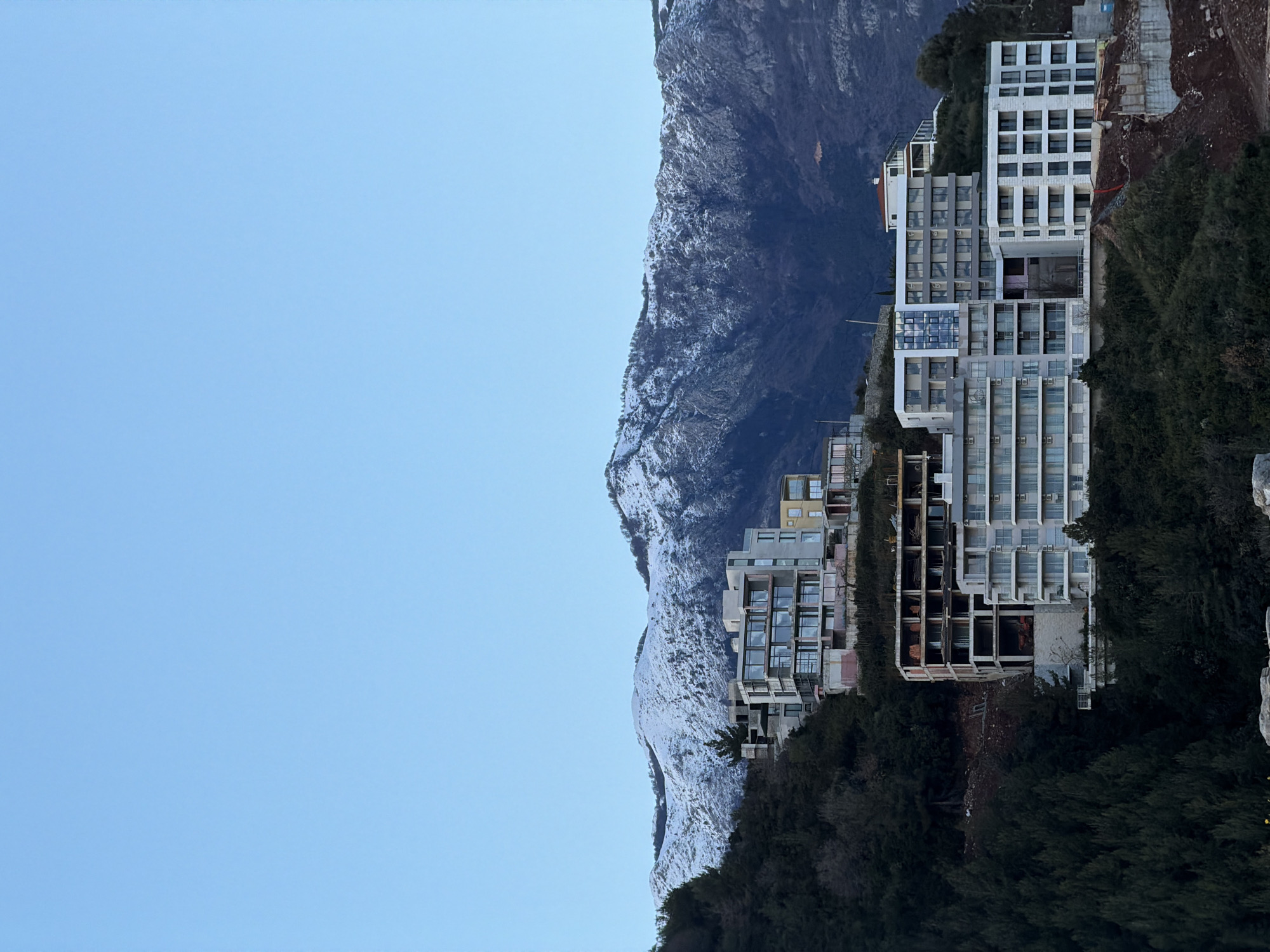 The image captures a mountainside scene in Budva, Montenegro. On the right of the image are tall, multi-story buildings built into the side of a steep hillside covered with dense green trees and shrubbery. The buildings appear to be either hotels or apartment complexes. To the left, a towering mountain range rises, its peaks partially covered with snow. The sky is a clear, pale blue, suggesting a sunny day.