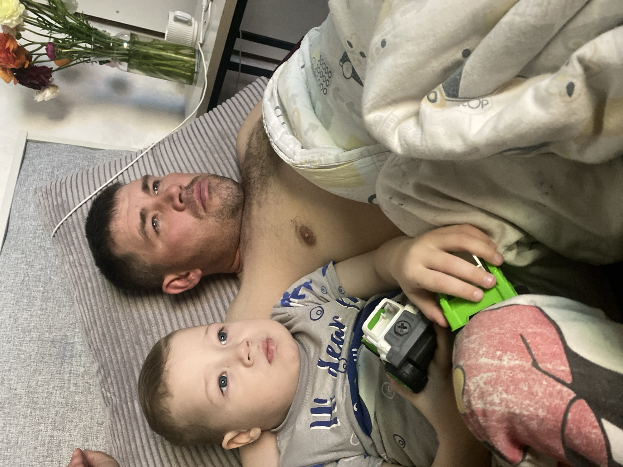 This image depicts an adult male and a young child (presumably father and son) relaxing together indoors, likely in a bedroom or living space. The setting is identified as Sochi, Russia. Both individuals are lying on their backs on a grey, striped mattress or pillow, looking upwards towards the camera.
The adult male, with dark hair and a short stubble beard, is partially shirtless. Next to him, the young boy, with light hair and blue eyes, is wearing a grey t-shirt with blue patterns and the partially visible text "My dear". He is holding a green and white toy truck in his hands. A colorful blanket, adorned with cartoon patterns including bears, numbers, and tools, covers part of the child and drapes over the adult's side.
In the background, on what appears to be a bedside table or shelf, a clear vase holds a bouquet of various flowers, including shades of red, orange, white, and pink. A white electronic device, possibly a charger or small appliance, is also visible on the surface, connected by a white cord.
The scene conveys a peaceful and relaxed moment, suggesting a quiet time together, perhaps in the morning or evening. Both subjects have calm, somewhat contemplative expressions. The photograph is taken from a slightly angled, top-down perspective. No specific weather conditions are discernible from the indoor setting.