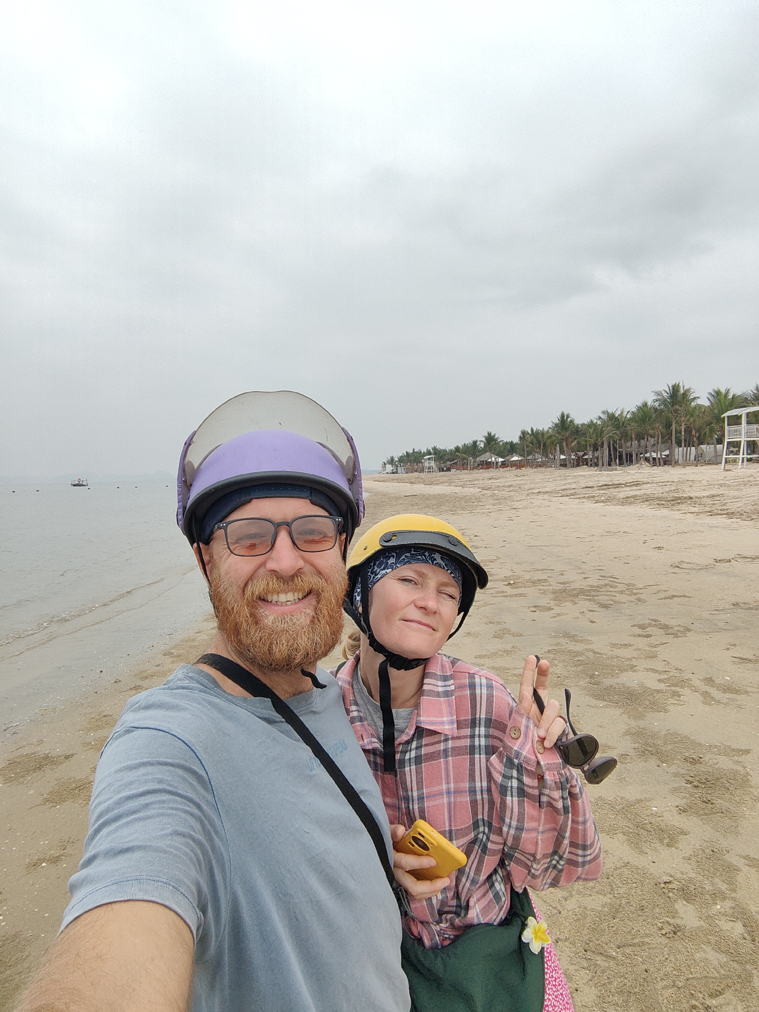 A selfie of two people on a beach. A man with a red beard wearing a blue shirt and a purple helmet is smiling. A woman wearing a yellow helmet and a pink plaid shirt is holding sunglasses and giving a peace sign. They are standing on a sandy beach in Bai Chay Ward, Vietnam. In the background, there is a body of water, palm trees, and structures that appear to be lifeguard towers. The sky is overcast.