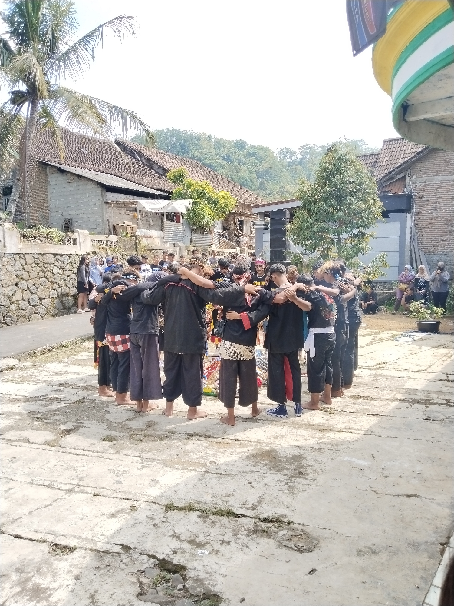 A group of people are in a circle with their arms around each other. They are dressed in black tops and pants, some with patterned wraps around their waists. They are standing on a cracked concrete surface. In the background are houses, trees, and a hillside. A crowd of people is watching. A large palm tree is visible on the left side of the frame.