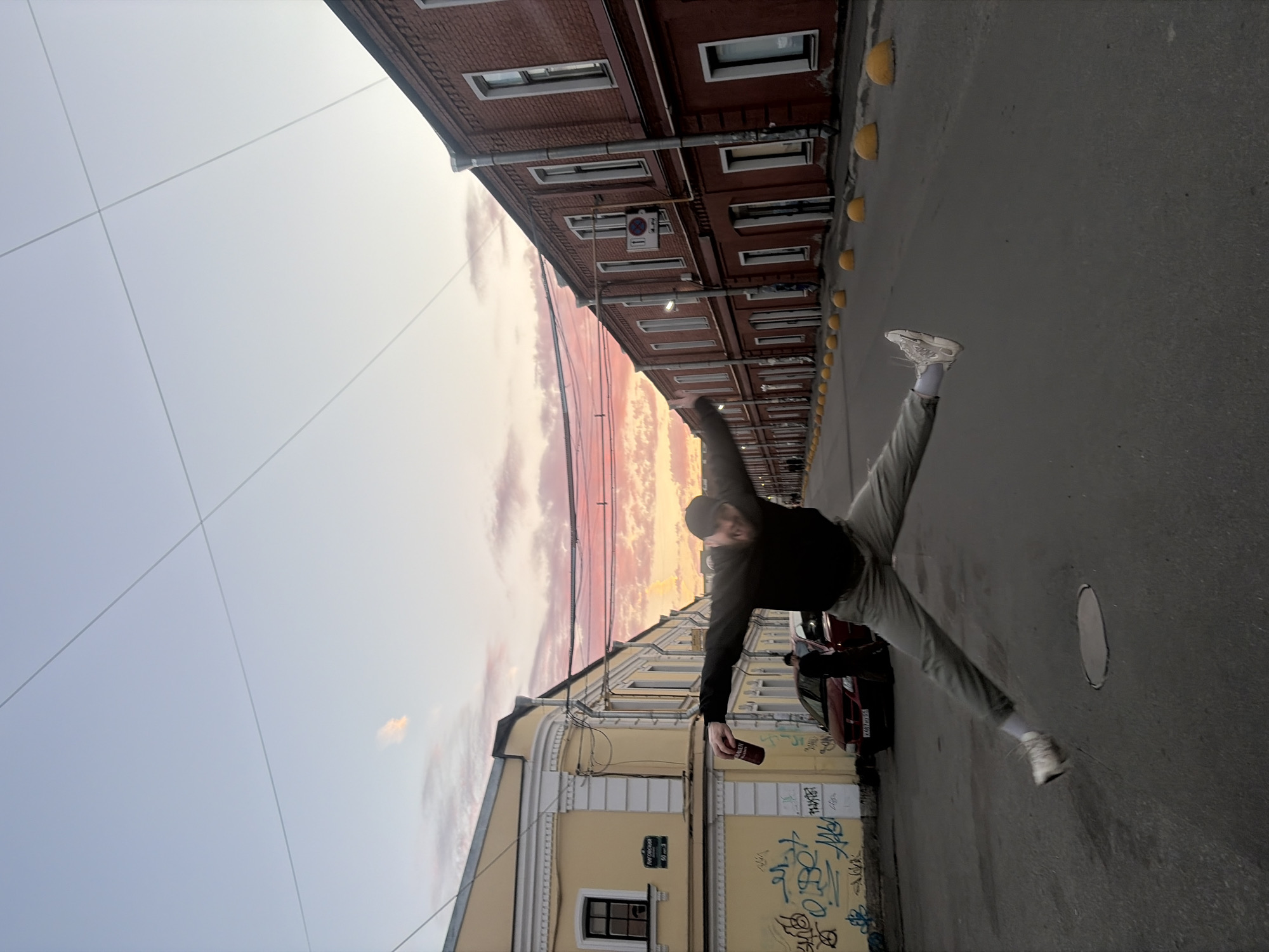 The image depicts a man mid-air, performing a jump or split with his legs extended in opposite directions. He is wearing a dark hooded sweatshirt, light-colored pants, and white sneakers. In his right hand, he holds a can. The background features an urban street scene with buildings on both sides. The building on the left is yellow with white trim and graffiti on its lower facade. The building on the right is made of red brick. A red car is parked on the left side of the street, and other pedestrians are visible in the distance. The sky above displays a gradient of colors, indicating a sunset or sunrise, with hues of pink and orange. The street is paved, and there are yellow bollards lining the right side. The overall impression is a candid moment captured in an urban environment during twilight. The street sign visible on the yellow building reads "Рогожская 50м" which translates to "Rogozhskaya 50m." Rogozhskaya Street is located in the city of Saint Petersburg, Russia.