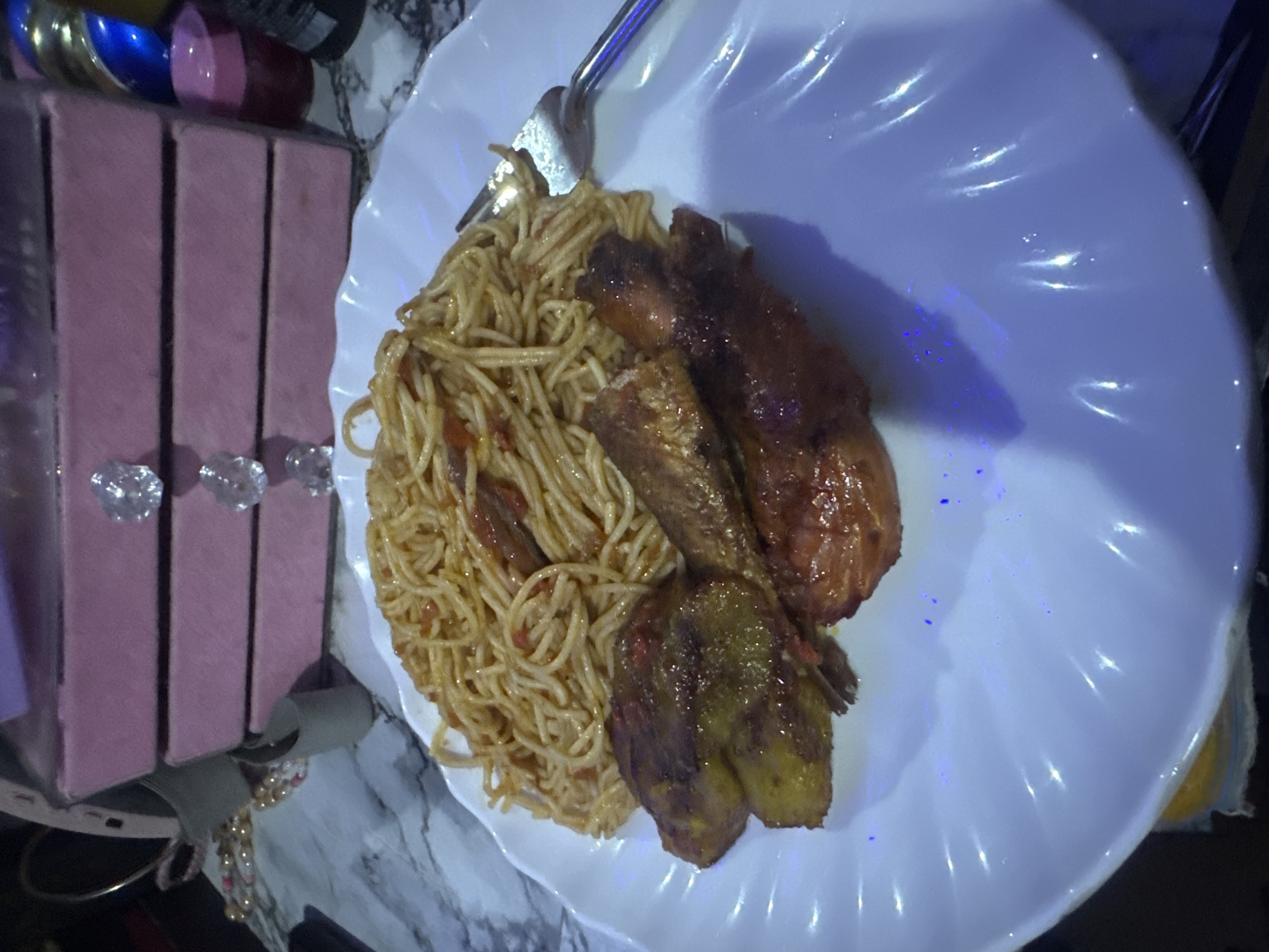 A close-up, overhead shot reveals a meal served on a white, wavy-edged plate, resting on a marble-patterned surface. The main subjects are a portion of spaghetti mixed with a light, possibly tomato-based sauce, and several pieces of what appear to be fried or roasted plantain and chicken, all with a dark, glossy, golden-brown finish. A fork or spoon is partially visible at the top of the plate, resting on the spaghetti.
In the background, to the left, are three stacked, rectangular boxes or containers in a muted pink or mauve color. Each box features a clear, diamond-shaped handle or knob. Behind these boxes, there are glimpses of other metallic or reflective objects, including blue, gold, and maroon items. A string of light-colored beads is also visible near the bottom left, resting on the marble surface.
The scene is indoors, likely a dining or kitchen setting. The lighting is somewhat dim, with prominent blue and purple reflections visible on the white plate, possibly from artificial light sources. No people are visible, and no text is discernible in the image. The food presentation suggests a prepared meal.