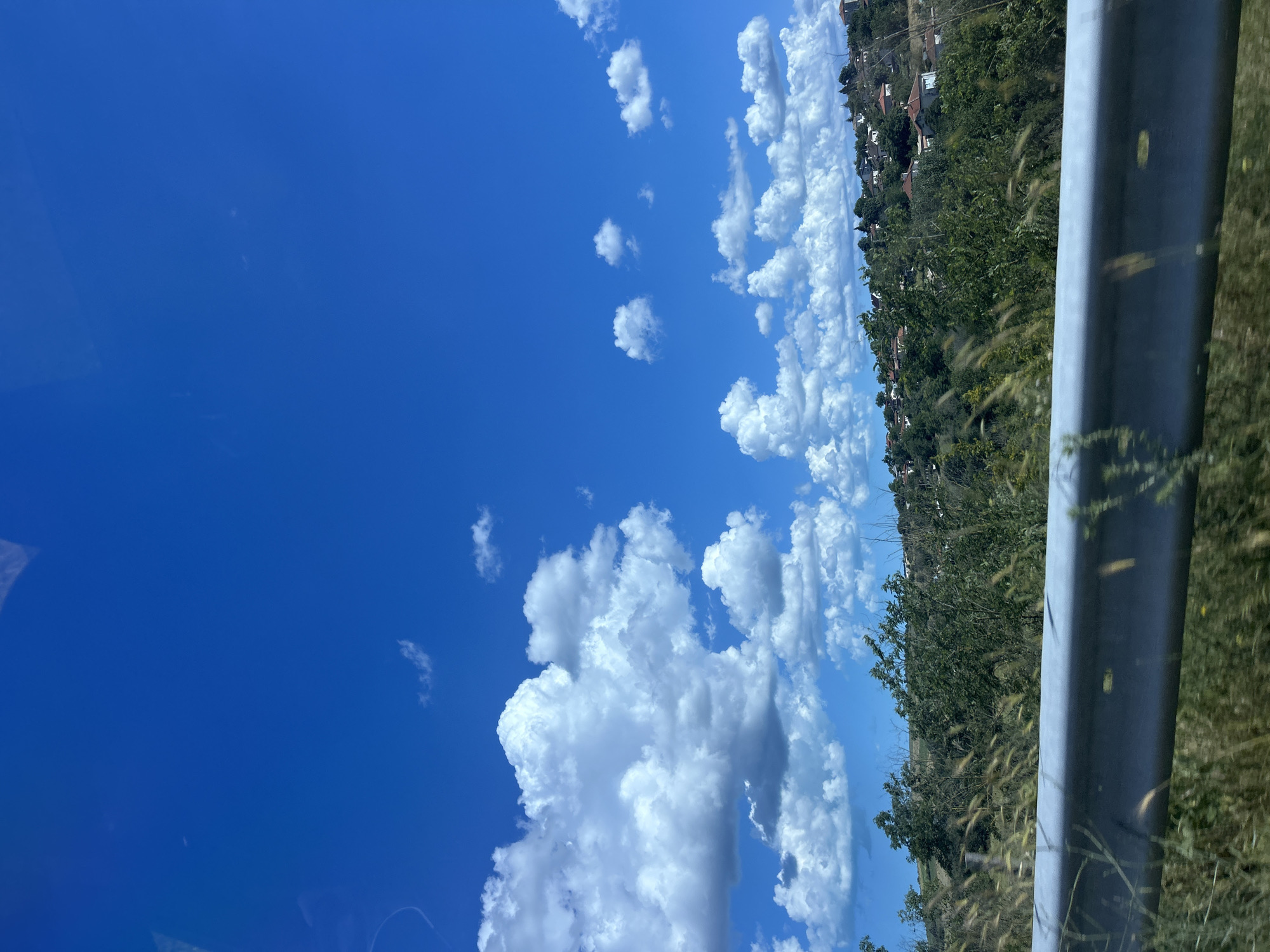 The image depicts a scenic view of a hillside village under a bright blue sky dotted with fluffy white clouds. The foreground features a grey metallic guardrail running horizontally across the frame, with dry grass and vegetation blurred in front of it. Behind the guardrail, a lush green hillside is covered with trees and bushes. Scattered across the hillside are numerous houses with red-tiled roofs, characteristic of the architecture found in Konstantinovo, Bulgaria. The scene suggests a rural or semi-rural setting on a clear day.