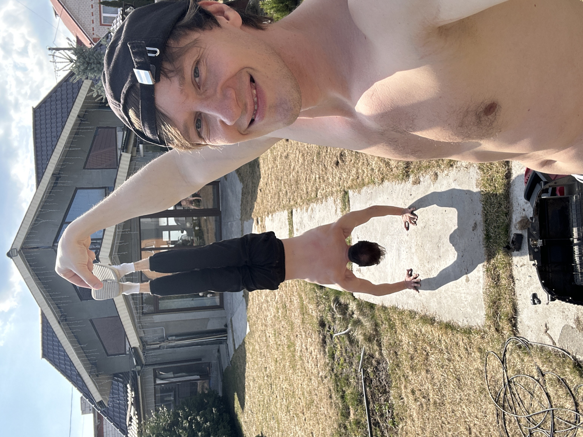 A shirtless man wearing a black cap is taking a selfie while holding the feet of another shirtless man, who is balancing upside down on his hands on a concrete path. The second man is wearing black pants and white socks. The background includes a house with a dark gray roof, a lawn of dry grass, and a high-pressure washer with hoses. The city of Kursk, Russia, is not discernible in this image.
