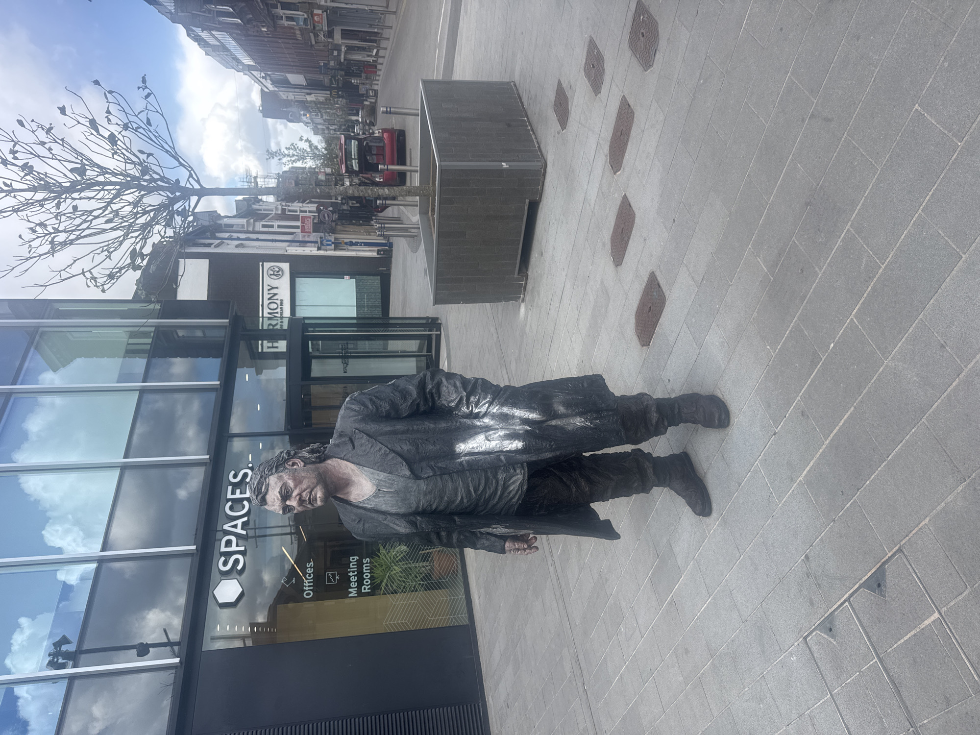 This image captures a pedestrianized urban scene in Woking, United Kingdom, specifically near Block E, The Lightbox, Dukes Walk.
In the foreground, a male street performer, or "living statue," stands facing towards the viewer's right. He is covered head-to-toe in metallic grey-bronze body paint, including his face, hands, and clothing. He wears a long, dark overcoat, a dark t-shirt, and dark trousers, all appearing to be painted to mimic aged metal or stone. Visible white paint streaks are present on his left arm and the front of his overcoat. His expression is neutral.
To the left of the performer is a modern building with a dark facade and large, reflective glass windows that reflect a partly cloudy sky. The ground floor displays a prominent white "SPACES." logo, with smaller "Offices" and "Meeting Rooms" text below. A revolving glass door serves as an entrance.
To the right of the performer, and extending into the background, is a wide, paved street. A bare tree, featuring dark, sparse branches and small, dark ornamental leaves, is planted within a large, dark grey rectangular planter. Further down the street, older brick and stone buildings line both sides, one displaying a "HARMONY" sign. A red vehicle with an open tailgate is parked in the distance. The ground is paved with light grey rectangular flagstones, interspersed with various rectangular and square dark metal utility covers. The sky is partly cloudy.