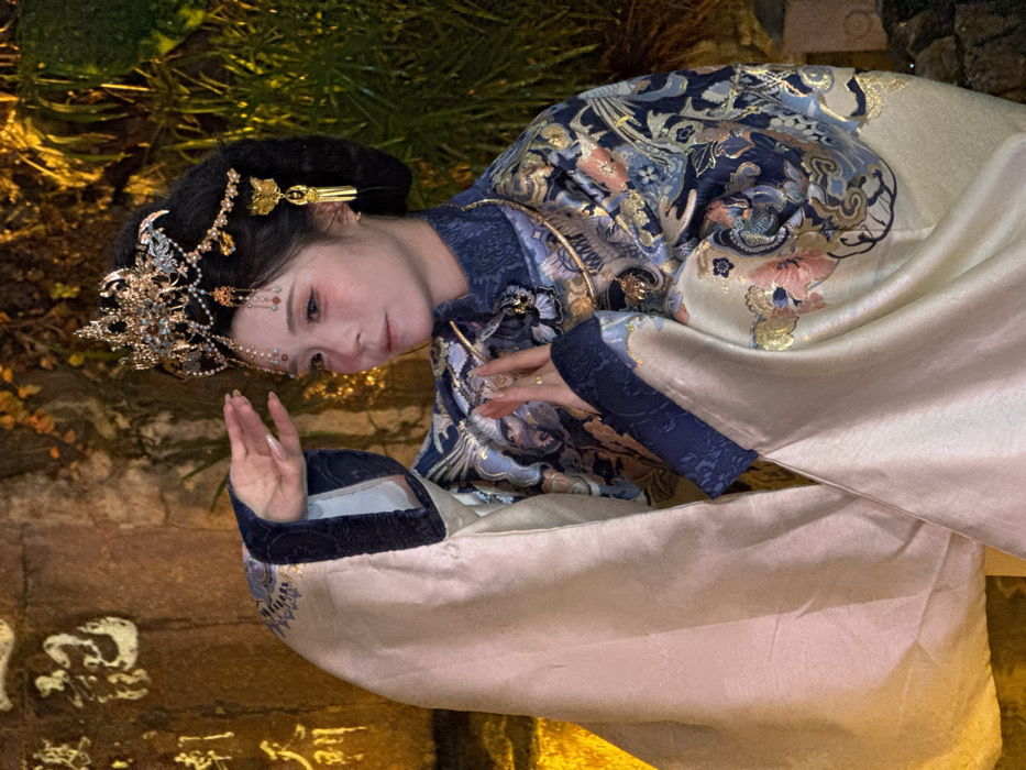A forensic examination of the image reveals a single East Asian female, positioned slightly off-center to the left, captured from the waist up. She is adorned in elaborate traditional Chinese attire, featuring an intricate navy and cream robe with gold, blue, and pink floral and patterned embroidery, overlaid with a flowing, light cream outer garment. A golden neck ring is visible beneath her high collar. Her dark hair is styled in an ornate updo, crowned with a large, golden headdress embellished with filigree, blue and red beads, and dangling chains. Golden, butterfly-like ear ornaments are also present. Her face is made up with blush and precise eyebrow definition. She holds her right arm raised with the palm open and fingers extended, while her left hand is posed closer to her chest with fingers slightly curled, displaying a gold ring.
The background consists of a warm, artificially lit environment, suggesting nighttime. To the left, a textured stone wall is visible, upon which Chinese characters are carved, appearing golden under the illumination. Partially visible characters include "朝天" (Chaotian) and "記" (Ji). To the right, lush green foliage is illuminated by similar warm lighting. The carved characters "朝天门" (Chaotianmen) on the stone wall strongly indicate the location as the Chaotianmen Square/Pier area in Yuzhong District, Chongqing, China.