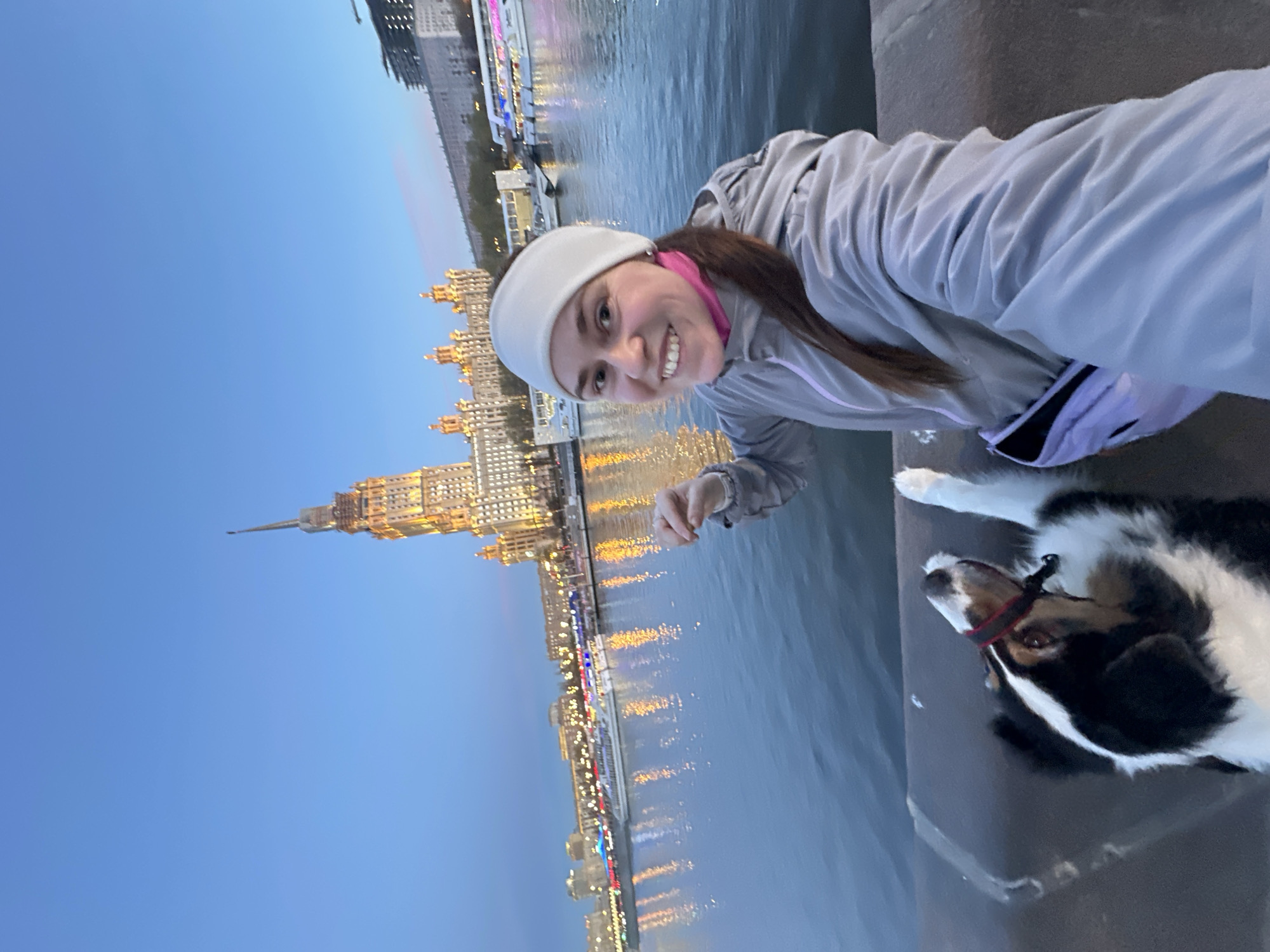 The image is a vertical selfie featuring a smiling young woman and her tricolor dog (black, white, and brown) against a backdrop of the Moskva River and illuminated city architecture at dusk in Moscow, Russia.
The woman, with long brown hair, wears a light grey athletic jacket over a pink top and a white headband. She is looking directly at the camera with a happy, content expression. Her dog, wearing a red collar, is lying down calmly next to her on a dark, stone-like surface, likely a river embankment.
In the background, a prominent Stalinist-era skyscraper, illuminated with golden lights, rises majestically, its spire piercing the deep blue twilight sky. This distinctive building is characteristic of Moscow's 'Seven Sisters.' Numerous other city lights along the riverbanks and from what appear to be boats are visible, reflecting brightly on the calm water. The scene captures a peaceful urban evening, highlighting leisure time with a pet against an iconic Moscow landmark. There is no visible text in the image.
