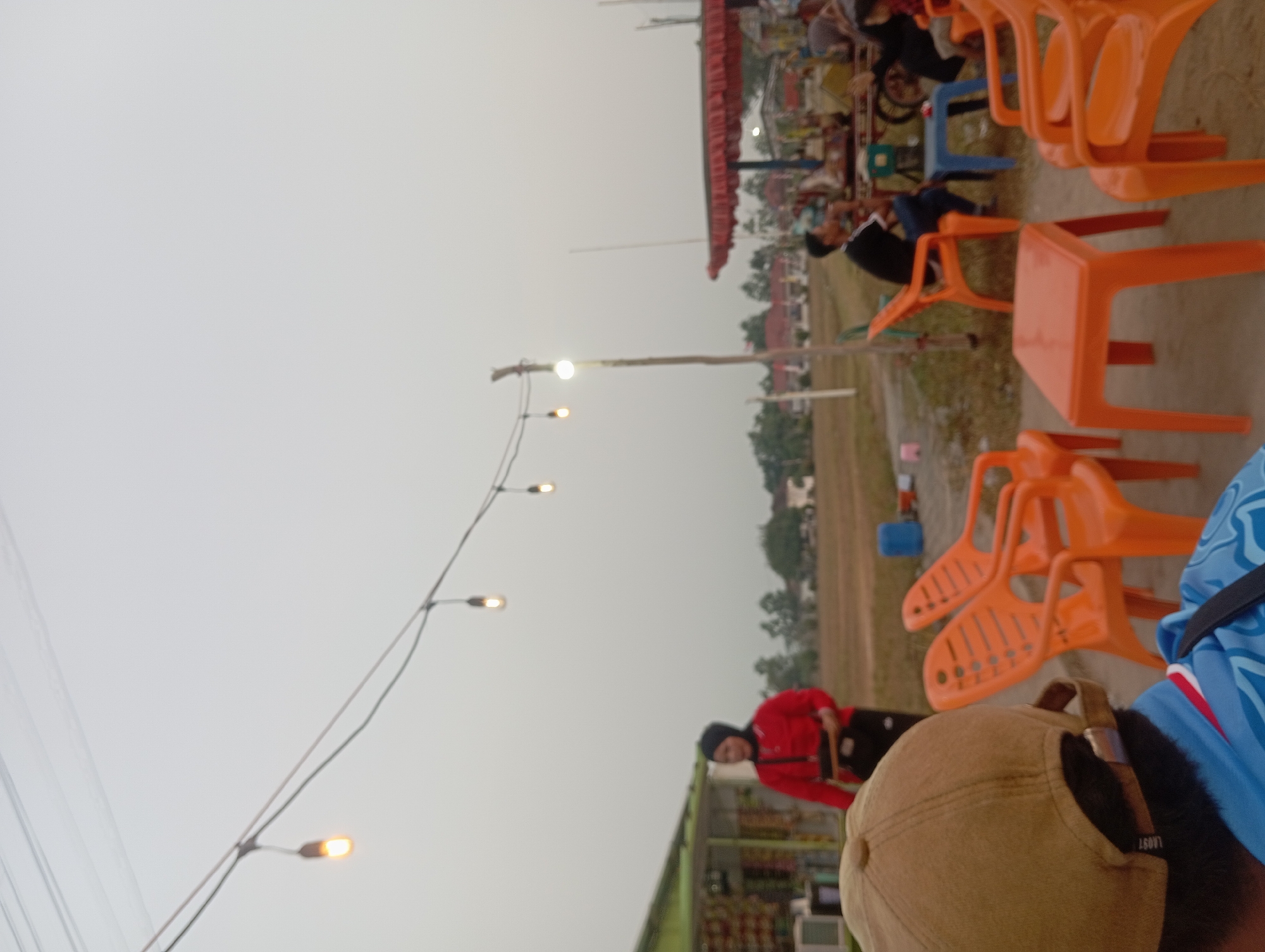 In Way Dente, Indonesia, at dusk, a small outdoor gathering takes place. A woman in a red shirt and black hijab stands by a roadside stall selling snacks. Several orange plastic chairs and tables are arranged in the open area, with a few people seated. One person wears a brown baseball cap and a blue shirt, facing away from the camera. Another person, wearing a black t-shirt, sits at a blue table. In the background, there are buildings and trees under a hazy, overcast sky. String lights are hung overhead, with a few bulbs illuminated. The ground is dry and dusty.