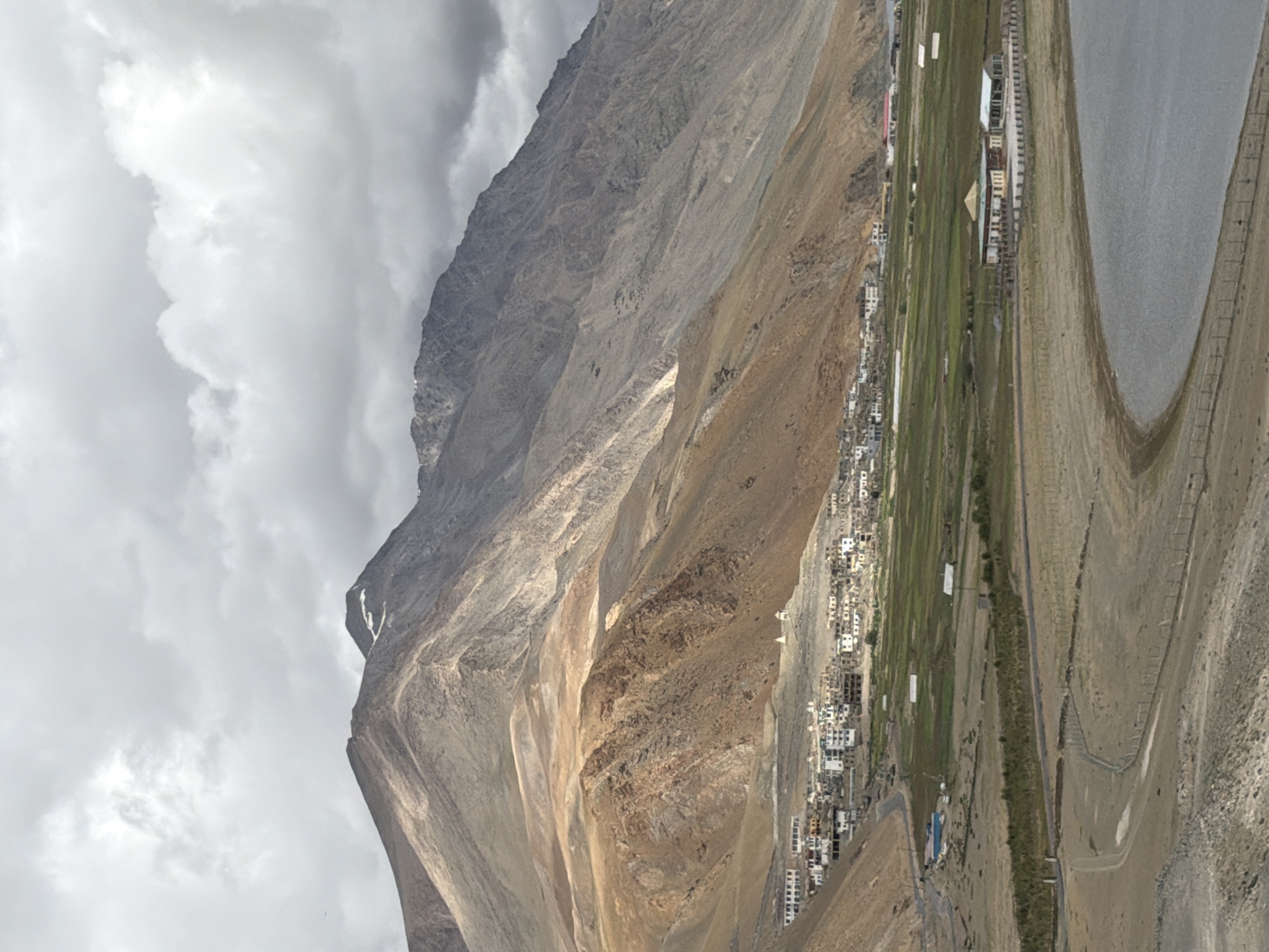 An aerial, distant, vertical, slightly overcast, wide shot captures a settlement nestled at the foot of a large, arid mountain range in Karzok, India. The mountain is a mix of tan, brown, and grey rock, with hints of snow on its upper peaks. In the mid-ground, a village of white buildings with flat roofs is clustered on the lower slopes of the mountain. A road winds through the village and leads to a patch of vibrant green fields. A body of water, appearing as a lake or river, occupies the foreground, its surface a muted blue-grey. The shoreline is sandy and bare, with a low fence running along a portion of it. A modern building with a central pagoda-like roof and an adjacent structure with a white roof sit near the water's edge. The sky is filled with heavy, grey clouds. There are no people visible in the scene.