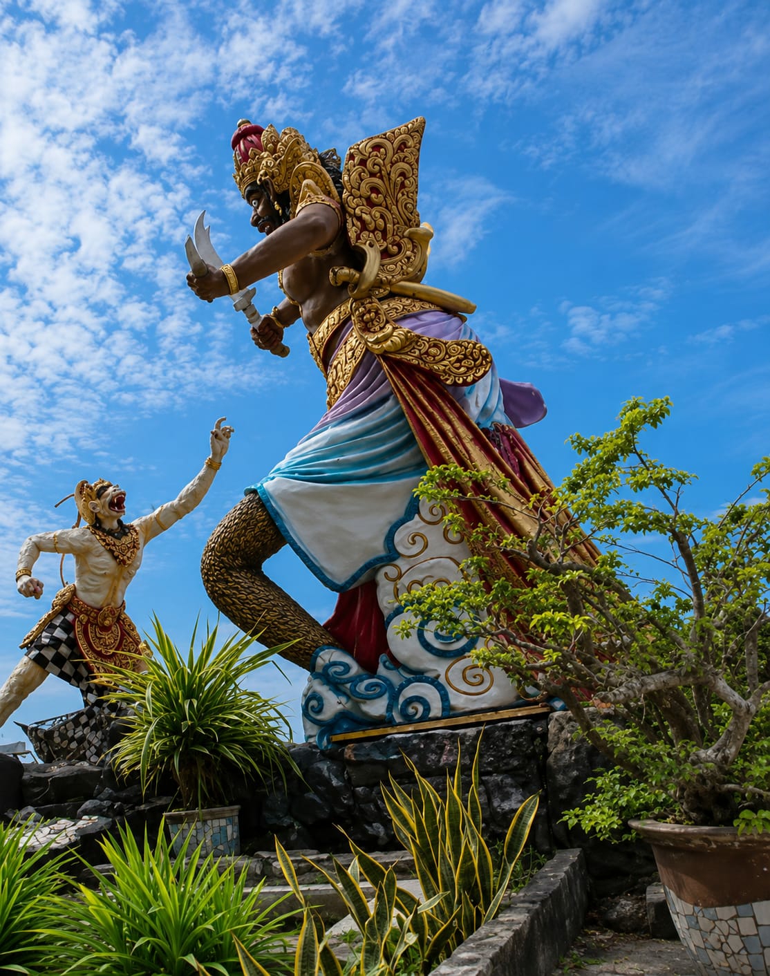 This is a low-angle, full shot of two imposing, colorful statues in Kota Pari, Indonesia, set against a bright blue sky with scattered white clouds. The statue on the right depicts a large, muscular figure with dark skin, adorned in ornate gold headdress and armor, a flowing sash of purple, blue, and white fabric, and red and gold draped cloth. It wields two curved swords. To the left, a smaller, light-skinned statue, resembling a monkey or mythical creature, with a human-like body, is depicted in a dynamic pose, reaching upwards with its left hand. This figure wears a gold necklace and a patterned checkered skirt. Both statues are positioned on a rocky, uneven terrain, with lush green plants and a potted tree in the foreground. The overall scene suggests a dramatic narrative or mythological representation.