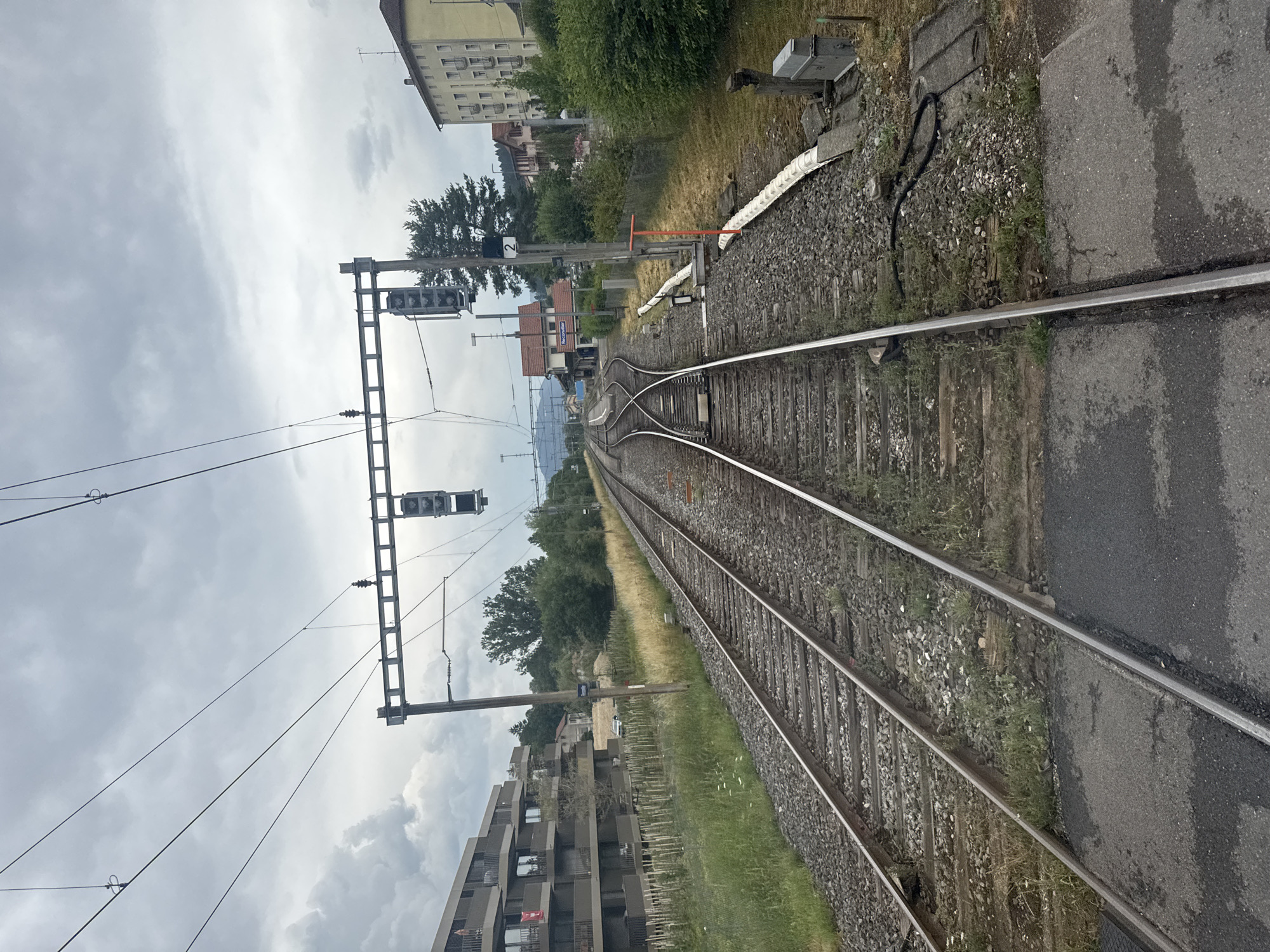 The image depicts a railway junction in Reconvilier, Switzerland, under a cloudy sky. Multiple sets of train tracks converge and diverge, with gravel and grass lining the ballast. Overhead power lines stretch across the scene, supported by metal gantries. A railway signal, displaying a vertical row of three lights, is visible on the left side of the image, with another similar signal on the right. In the background, apartment buildings and houses line the left and right sides of the tracks, respectively, interspersed with trees and vegetation. The scene appears to be a functional train station or a point of railway infrastructure. No people are visible in the image.