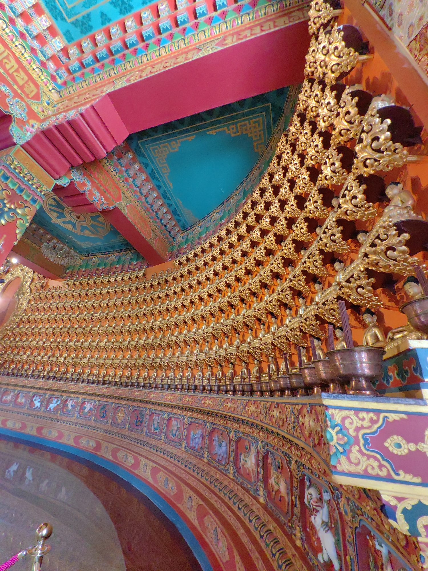 The image depicts the interior of a Buddhist temple, likely in Parsa, Nepal, given the provided context. The scene is characterized by ornate and vibrant decorations, showcasing a rich cultural and religious atmosphere. The ceiling is a riot of color, with intricate patterns in red, teal, and gold. Panels of geometric designs and stylized figures adorn the ceiling.

The main focus of the image is a curved wall lined with countless small, golden Buddha statues. These statues are arranged in rows, filling the entire visible surface of the wall. Each statue sits in a decorative golden holder, creating a mesmerizing, repetitive pattern. Below the rows of Buddha statues, the curved wall is further decorated with a frieze of painted panels. These panels depict various scenes and figures, rendered in a colorful and detailed style, suggesting narrative or devotional imagery. The colors used in this frieze include reds, blues, and ochres, with intricate borders and details.

In the foreground, a portion of the temple's architecture is visible, including curved surfaces and decorative elements. There are no people clearly visible in the image, suggesting a moment of quiet contemplation or that the photograph was taken at a time when the space was empty. The lighting appears to be artificial, highlighting the gold of the statues and the vibrancy of the painted surfaces. The overall impression is one of opulence, devotion, and artistic craftsmanship.