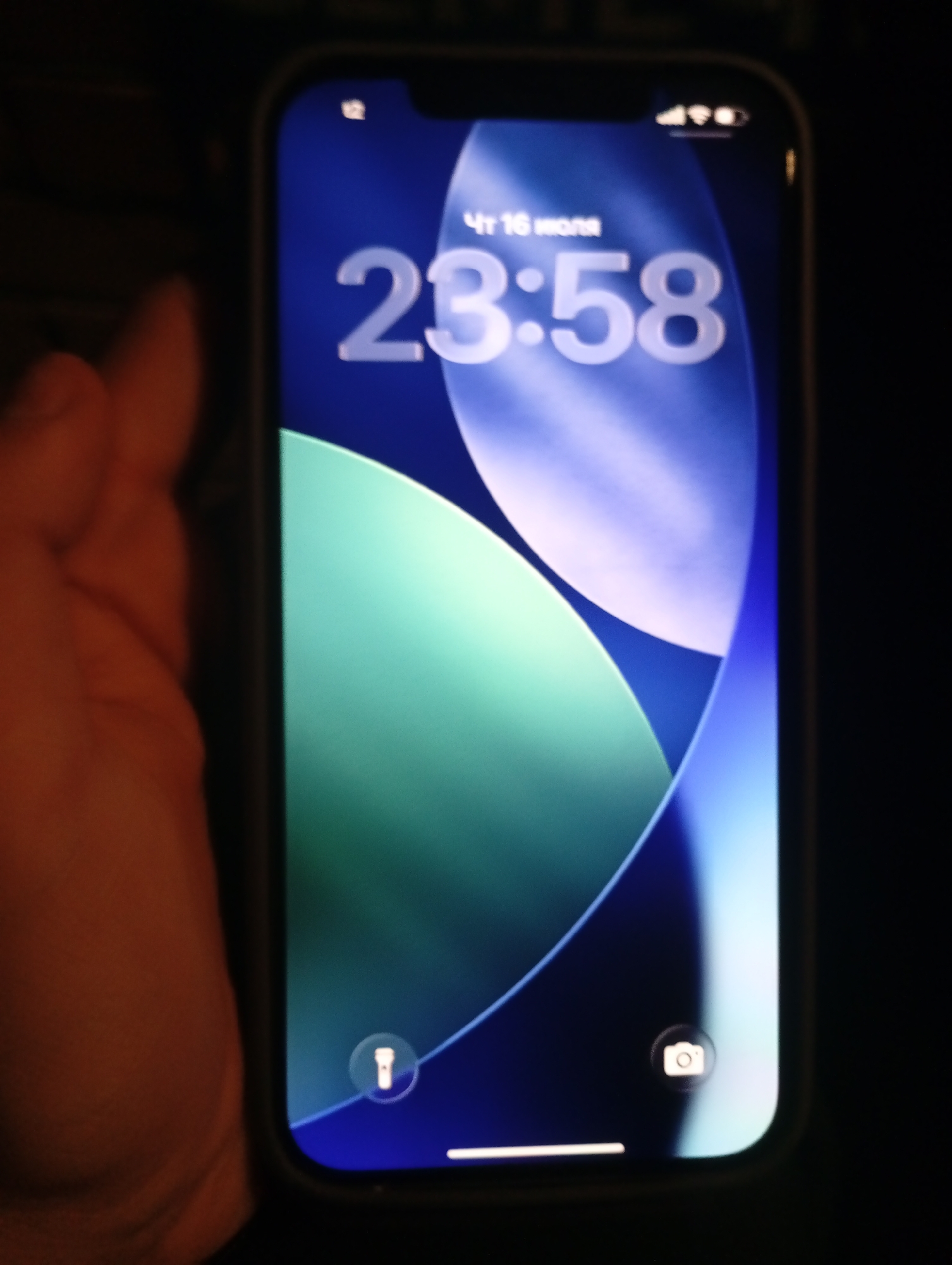 The image shows a close-up of an iPhone's lock screen. The time displayed is 23:58, and the date is July 16th. The wallpaper is a gradient of blue and green with flowing abstract shapes. At the bottom of the screen, there are icons for a flashlight on the left and a camera on the right. A hand is partially visible on the left side of the image, holding the phone. The background is dark, suggesting the photo was taken in low light conditions. There is no indication of the location being the city of Mayak, Russia.