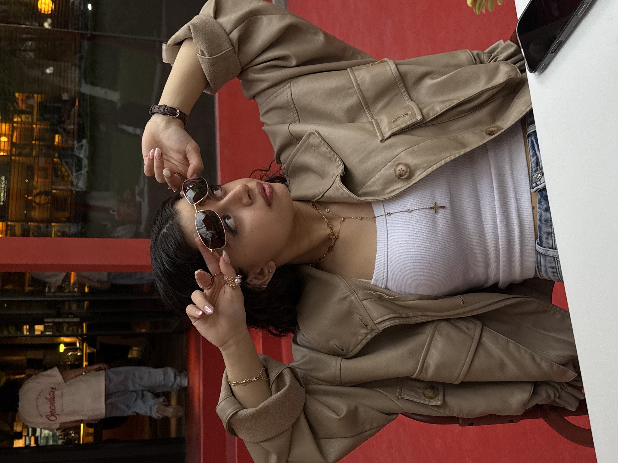 A young woman is sitting at a table outdoors, likely in Yerevan, Armenia, given the style of architecture and potential local fashion trends. She is wearing a light brown leather jacket over a white tank top, with blue jeans visible at her waist. She has sunglasses in her hand and is looking up and to her left. She is wearing a watch, multiple rings, and necklaces. A phone is visible on the white table next to her. The background shows a restaurant with people inside and red accents.