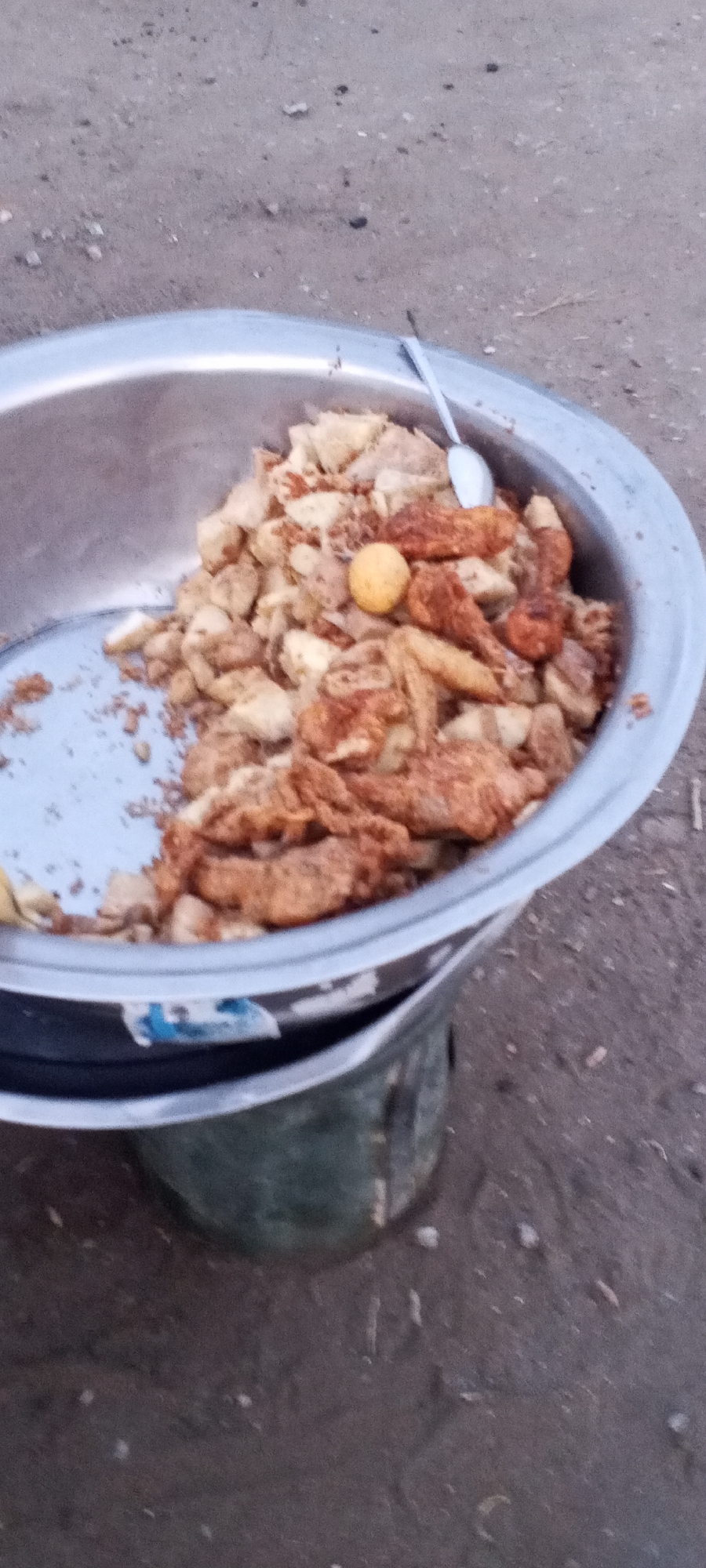 A metal bowl filled with food sits atop a container. The food appears to be a mix of fried items, including what could be chicken or similar breaded pieces, and what may be potatoes. There is also a single yellow item, possibly an egg or a fruit. A spoon rests on top of the food. The scene is set outdoors on a dirt surface. The location is indicated as Zango-ndaji, Nigeria.
