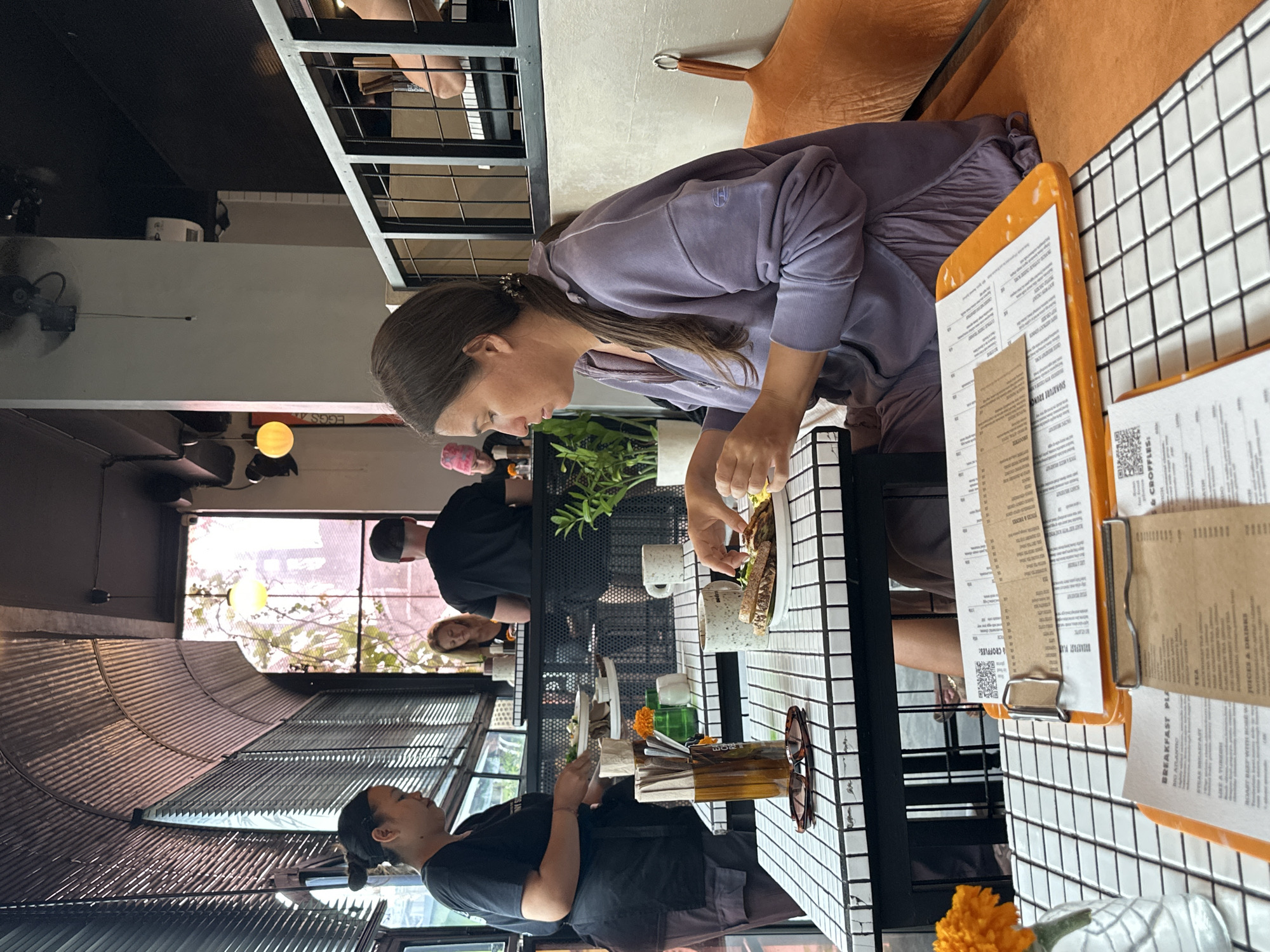 This image captures a moment at a cafe in North Kuta, Indonesia, likely during the daytime. Several people are present, engaged in dining or working. In the foreground, a woman with brown hair pulled back is seated at a table, reaching for food on a plate. She is wearing a lavender hoodie. The table is covered with a black and white tiled surface and holds plates, cups, and menus. One menu, propped on an orange clipboard, displays text that includes "BREAKFAST PLA," "SIGNATURE BRUNC," and "EGGS 4."
Behind her, another person in a black shirt and cap is visible, facing away from the camera. Further back, a woman with blonde hair appears to be looking towards the window. The cafe setting features large windows that allow natural light to enter, revealing greenery outside. Industrial-style metal accents are visible in the background, contributing to the cafe's aesthetic. Another person, a woman with dark hair tied up, is seated at a different table, also looking away. The overall atmosphere appears relaxed and casual, suggesting a typical cafe scene.
