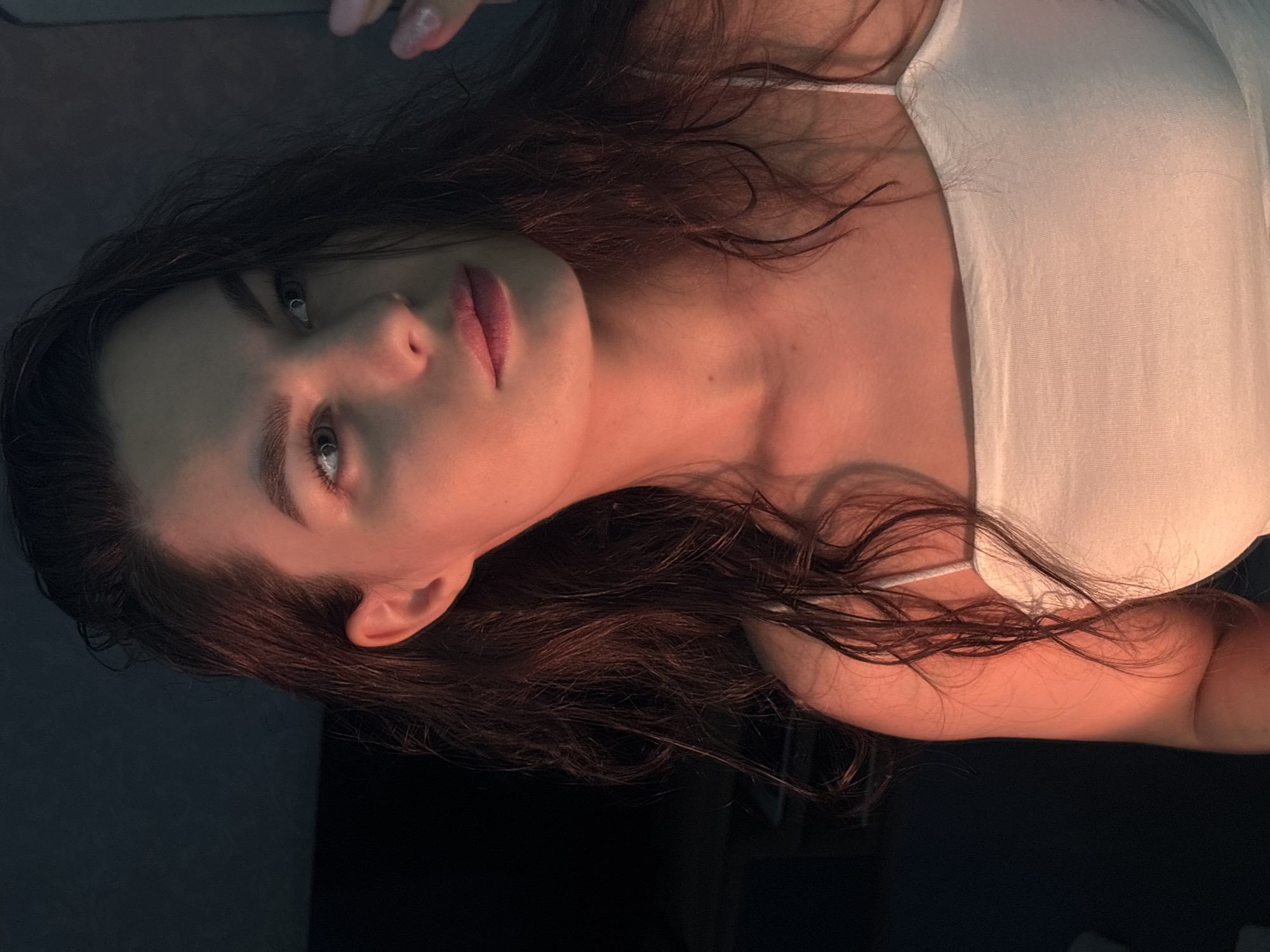 A young woman with long, dark, wavy hair and fair skin is captured in a close-up, head-and-shoulders shot. She is wearing a white tank top. Her head is turned to the right, and she is looking away from the camera with a pensive expression. Her left hand is raised, with fingers extended, near her right temple. The lighting is dramatic, with a warm, orange glow illuminating the right side of her face and upper body, while the left side and background are cast in shadow. The background appears to be an indoor setting with a dark wall. The image does not contain enough information to determine the specific location within Chaikovsky, Russia.