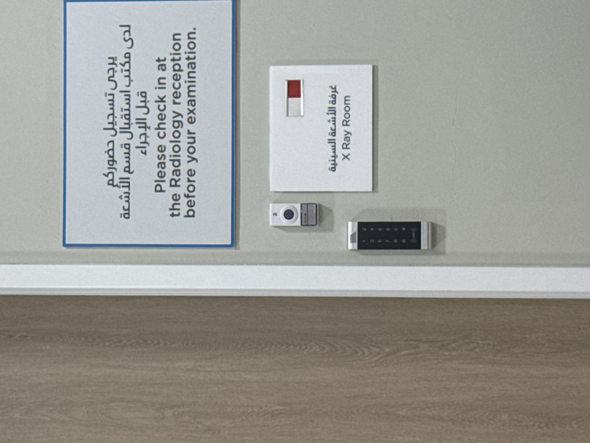 The image shows a wall in a medical facility. Two signs are displayed, one in both Arabic and English instructing patients to check in at the radiology reception before their examination, and another also in Arabic and English indicating "X Ray Room." Below the "X Ray Room" sign is a small camera and a keypad. The wall is painted in a neutral color, with a white baseboard running along the bottom. The floor is covered with wooden planks. The scene appears to be in Dubai, United Arab Emirates due to the presence of Arabic text alongside English on the signs.