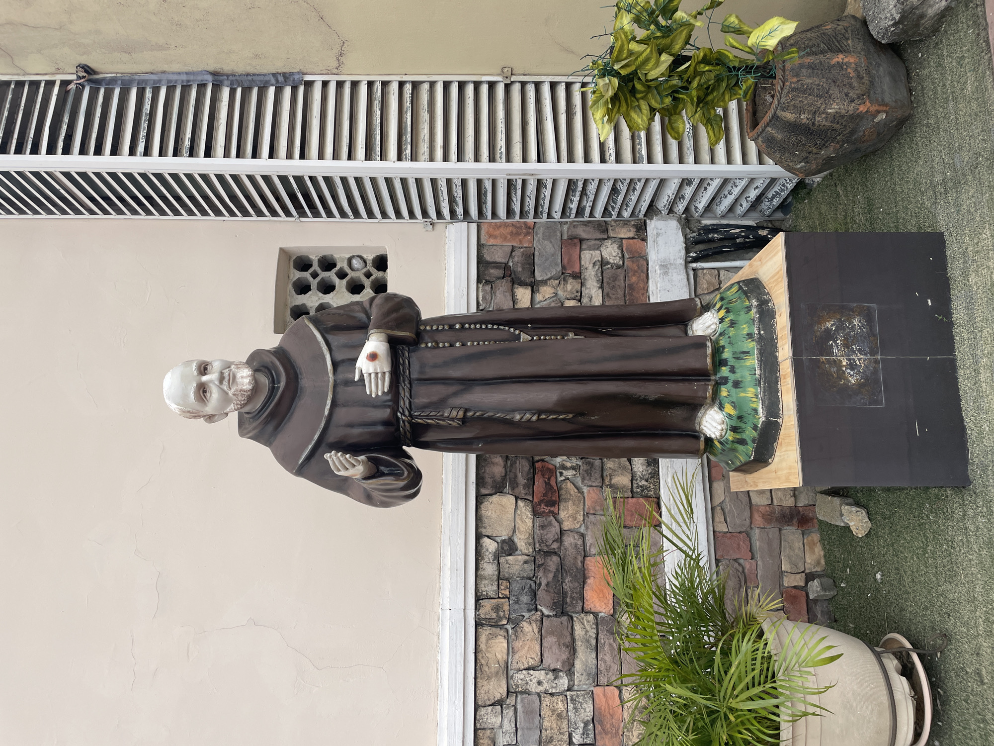 A statue of Saint Padre Pio stands outdoors in Unknown, Unknown. The statue is painted in brown robes and has a white beard and white hands. The statue is standing on a wooden and green base with a black top. The background includes a light-colored wall, a brick wall, a white vent, and potted plants. The environment appears to be a courtyard or garden.