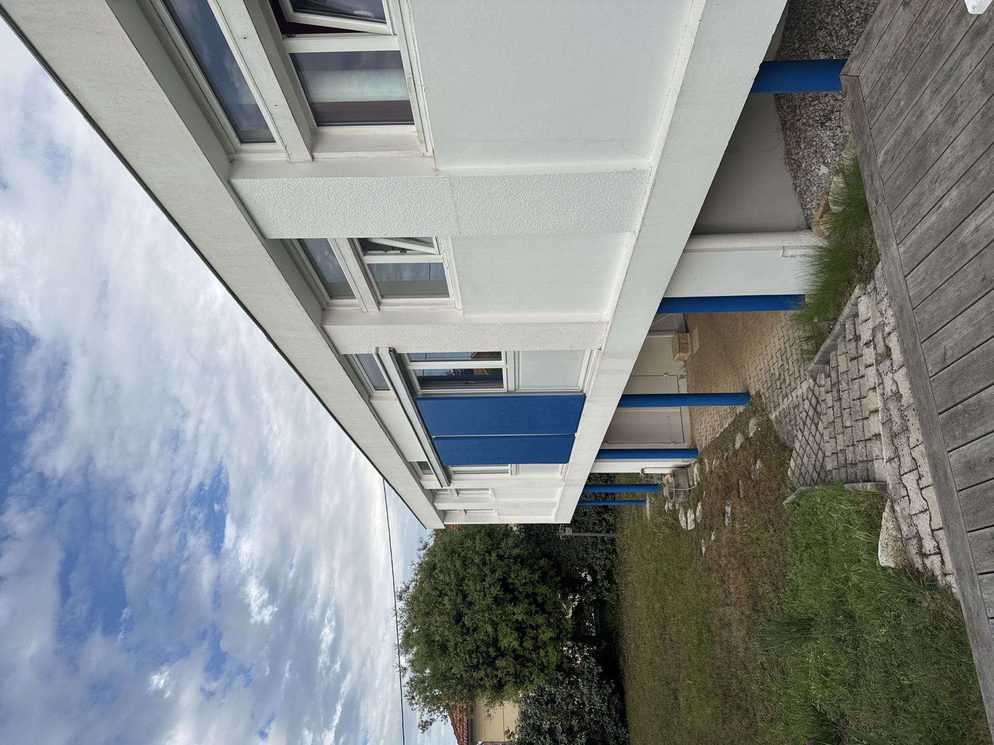A low-angle shot shows a modern building in Soulac-sur-Mer, France. The building features a white facade with windows, some of which are open, and sections painted blue. Blue cylindrical supports are visible beneath the building's overhang. A partly cloudy sky is visible in the upper left. The foreground features a grassy area with a tree and brickwork leading to a wooden deck.