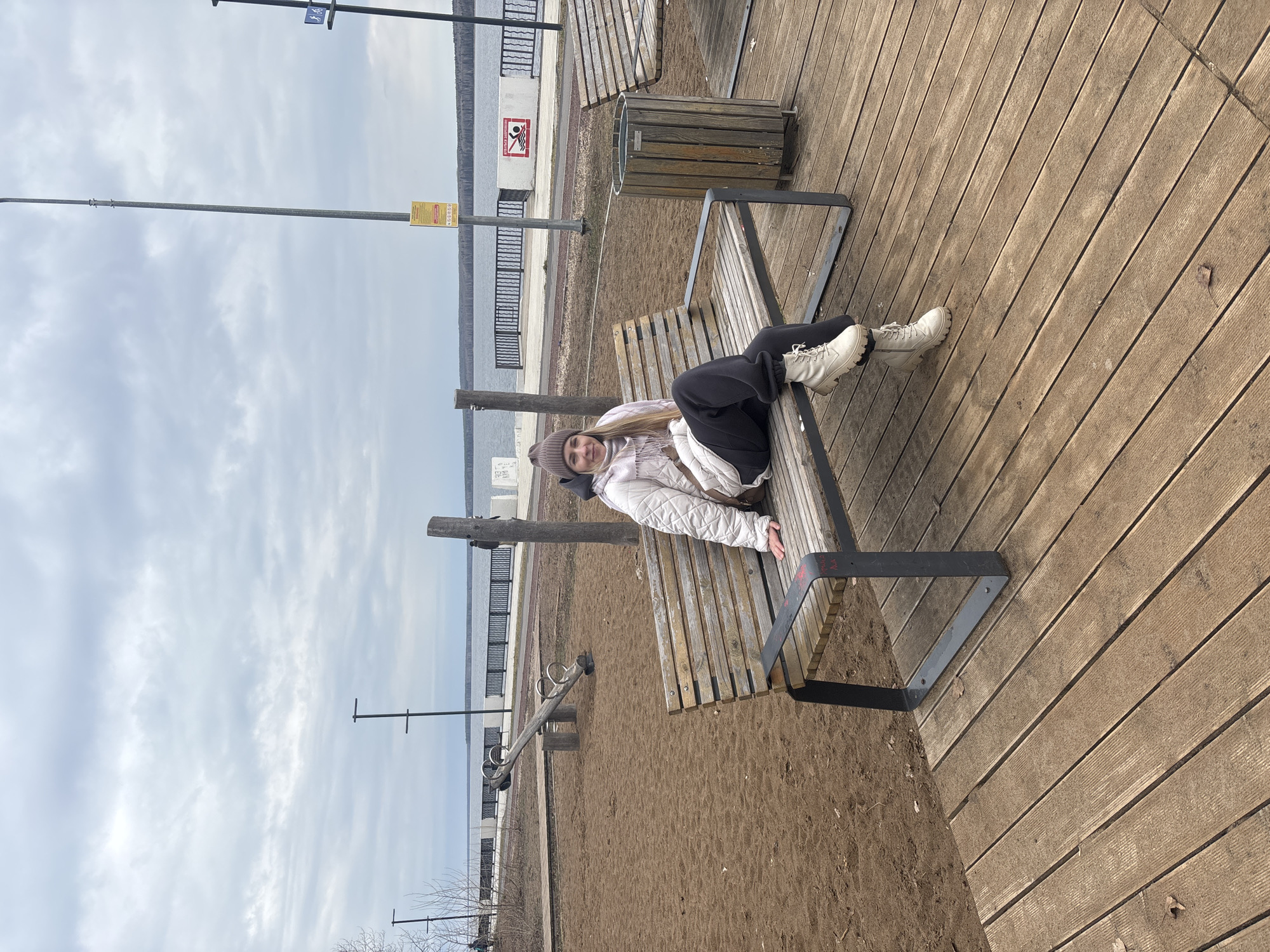 A woman is lying on a bench at a waterfront park. The woman is wearing a light-colored quilted jacket, dark pants, a beanie, and white boots. She is facing the camera with a slight smile. The bench is made of metal and wooden slats, and it sits on a wooden platform. In the background, there is a body of water, a walking area with railings, and the sky with clouds. A playground with a seesaw is on the sand. The picture was taken at a park in the city of Chaikovsky, Russia.