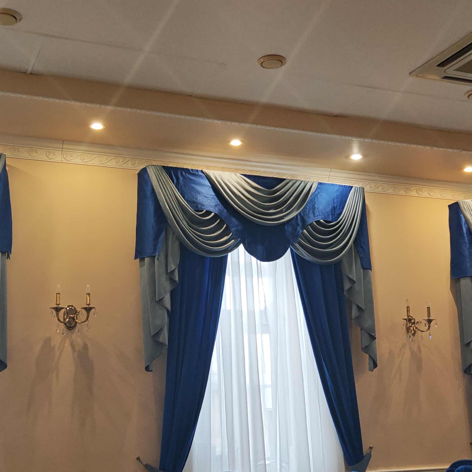 This image captures an interior scene, likely a formal room such as a dining hall or event space in Novo-Nikol'skoe, Russia. The primary subjects are the elaborate window treatments and the room's elegant decor.
A large window, covered by sheer white curtains, is flanked by ornate, two-tone drapes. The drapes feature a royal blue fabric combined with a lighter grey-blue, arranged in decorative swags and valances at the top, and flowing panels on the sides. Two antique-style, brass-colored wall sconces with crystal embellishments and electric candle bulbs are mounted on the light beige walls, one on each side of the central window. These sconces cast prominent, elongated shadows on the wall, suggesting illumination from the front.
The ceiling above is light-colored, possibly a suspended ceiling with acoustic tiles, and features several recessed spotlights that provide additional artificial illumination. A circular device, possibly a smoke detector, and an air conditioning vent are also visible on the ceiling. Decorative crown molding with a swirling pattern runs along the top of the walls, just below the ceiling.
The scene appears static with no people or ongoing activity. The artificial lighting and formal furnishings suggest an indoor setting designed for gatherings or events. No text is visible in the image.