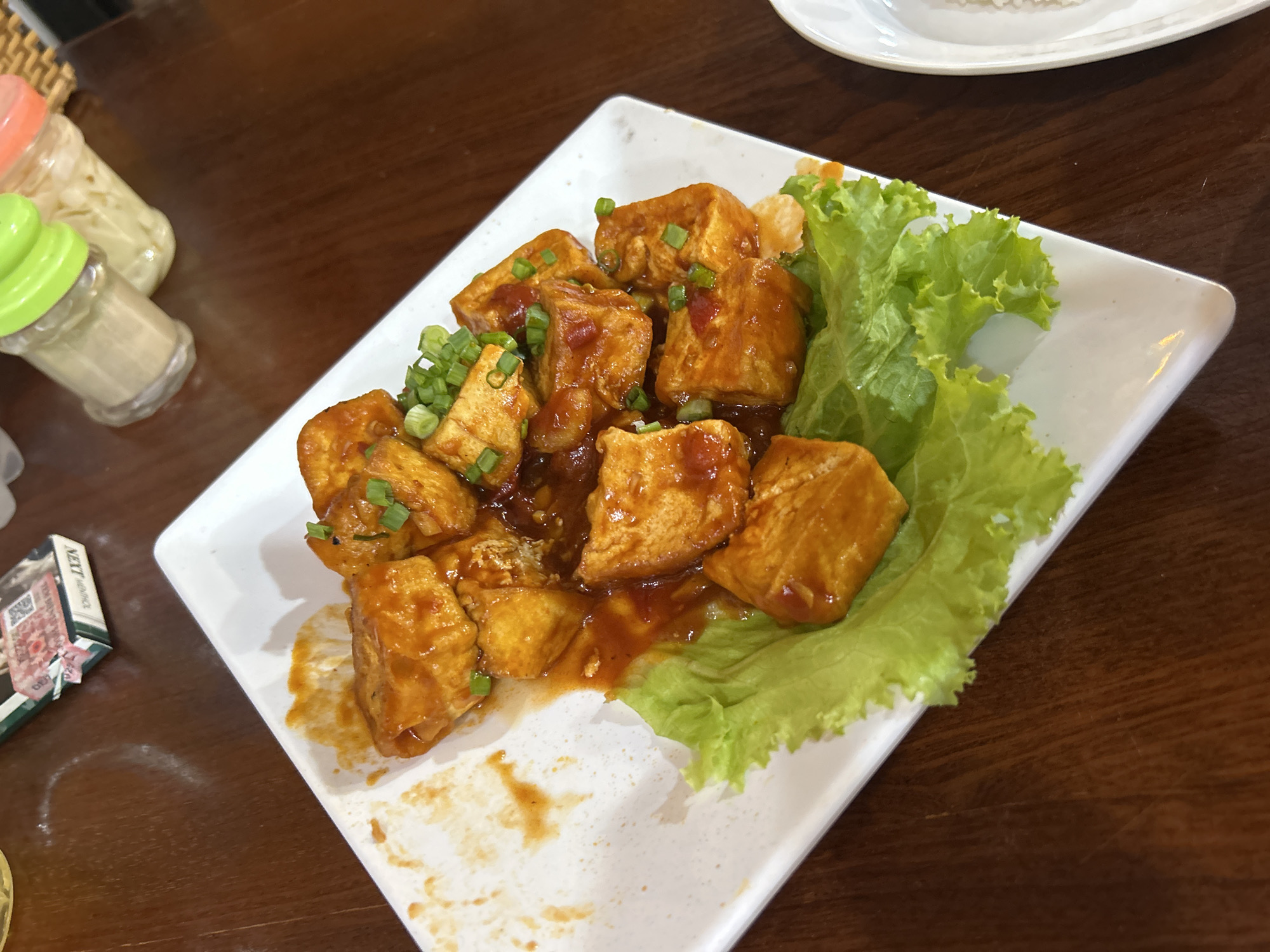 A square, white ceramic plate on a dark wooden table holds fried tofu cubes coated in a reddish-brown sauce, garnished with chopped green onions and accompanied by green lettuce leaves. To the left of the plate, there are two condiment dispensers, likely containing sauces or spices. A package of NEXT-branded cigarettes is also visible on the table near the plate. In the background, another plate, possibly containing rice, is present. The scene appears to be a casual dining setting. The city of Nha Trang, Vietnam, is specified as the location.