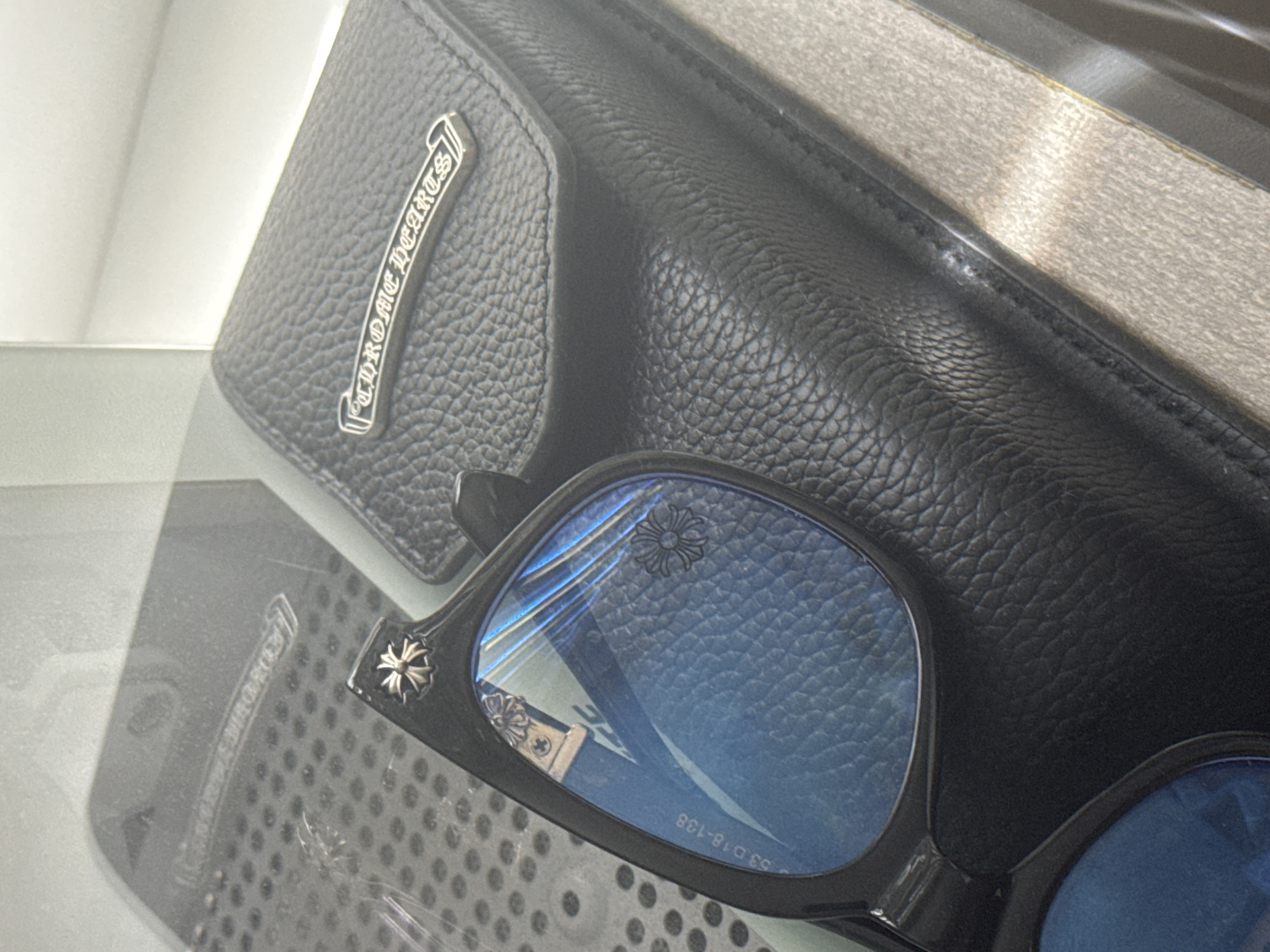 The image shows a close-up of Chrome Hearts sunglasses and a black leather wallet, possibly a Chrome Hearts wallet based on the branding on the wallet. The sunglasses have a black frame and blue-tinted lenses, with a Chrome Hearts logo visible on the temple. The wallet appears to be new and has a textured surface. The items are located in the city of Burgas, Bulgaria. The image captures a sense of luxury and fashion.