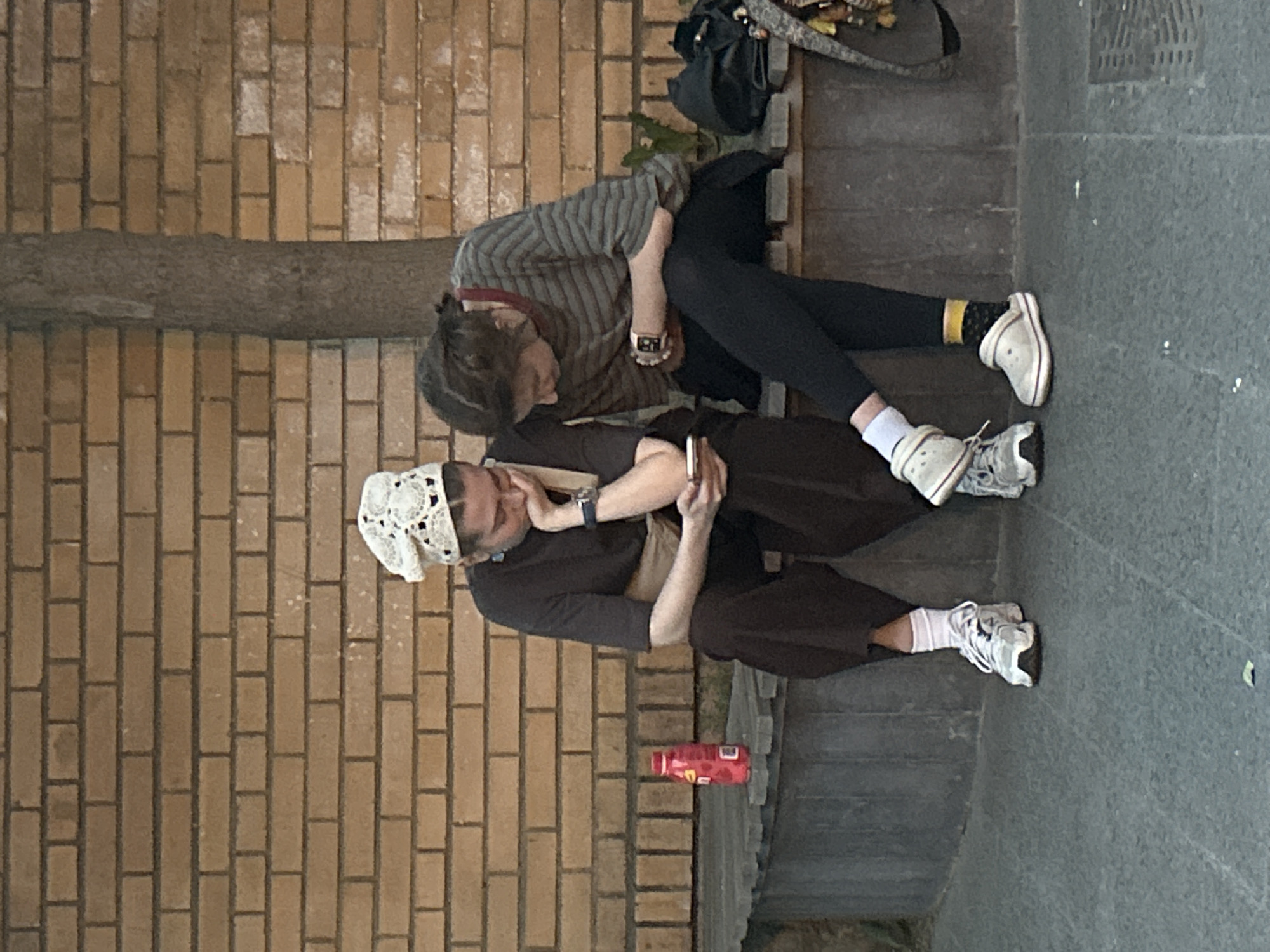 The image captures two individuals sitting on a set of concrete steps in an urban environment. A brick wall is adjacent to the steps. One person wears a striped shirt, dark pants, white socks with a yellow band, and white sneakers. The individual is leaning down, their face obscured. Next to them, a black backpack rests on the step. The second individual wears a dark shirt, loose brown pants, white socks, white sneakers, and a white lacy head covering. They appear to be looking at a mobile phone in their hands. A red bottle is nearby. The city is Unknown, Unknown.