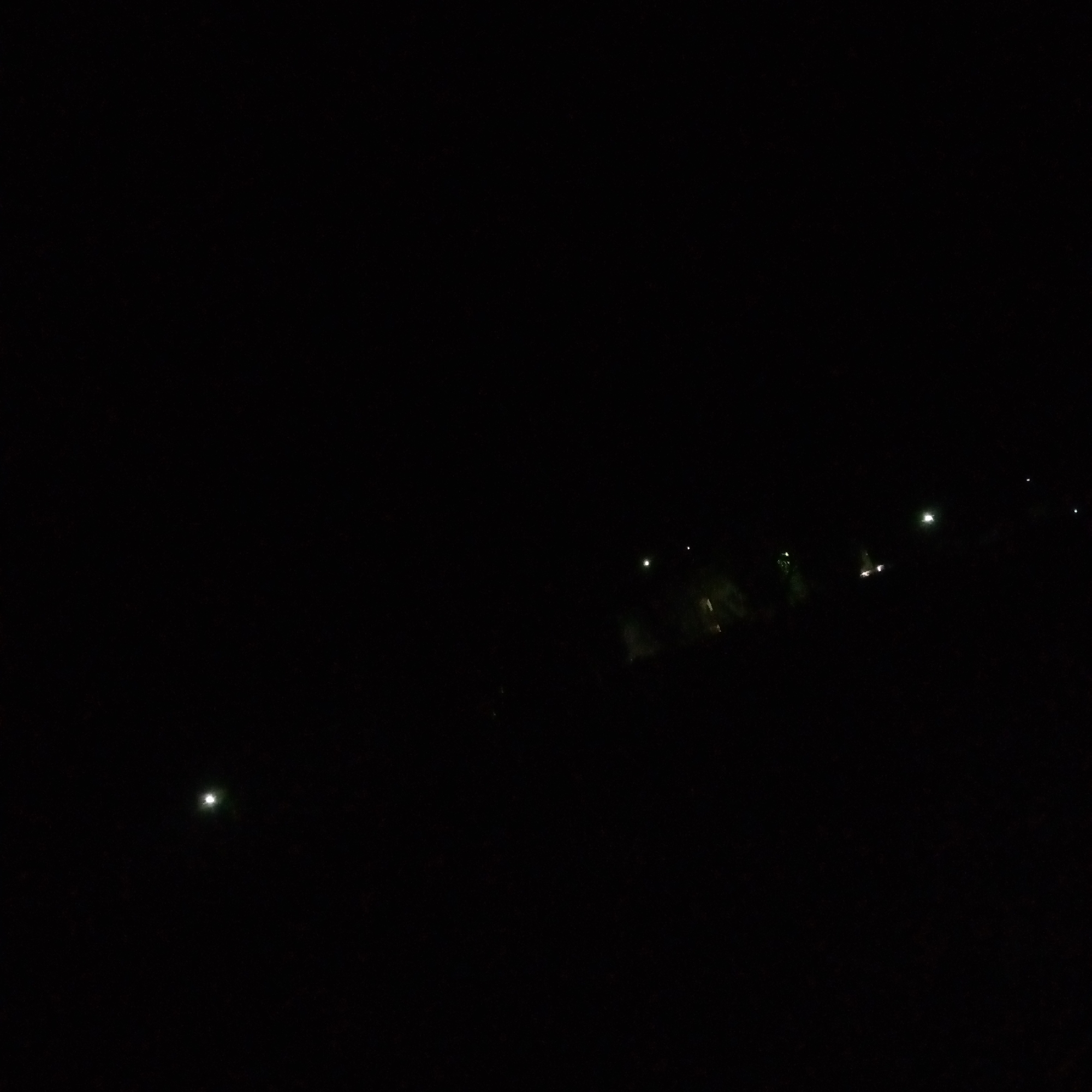A low-light, nighttime photograph captures a dark landscape dotted with distant lights. The scene is predominantly black, suggesting it's a night shot with minimal ambient light. In the lower left quadrant of the image, a single bright, round light source is visible against the darkness. Further to the right and slightly higher, a cluster of fainter lights appears, some appearing to be streetlights or building lights. The background is uniformly dark, hinting at a rural or undeveloped area rather than a densely populated urban center, or possibly a landscape with very little light pollution.