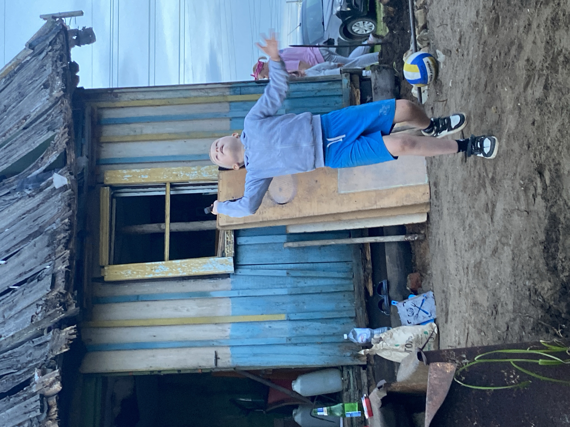 The image shows a young boy in mid-air, appearing to be jumping or performing a trick. He is wearing a light grey hooded jacket and blue shorts, with black and white sneakers. His arms are outstretched, and his eyes are looking upwards. To the left of the boy is a dilapidated wooden structure with weathered, grey wooden planks and blue and yellow painted sections. There is an open window with a wooden frame. In the background, a silver car is visible, and another person, possibly a girl, wearing a pink top and grey pants is lying down. A volleyball, colored blue, yellow, and white, rests on the sandy ground near the boy's feet. The ground is mostly dirt and sand. The overall scene depicts an outdoor, possibly rural or semi-abandoned setting. The sky appears overcast. The image is rotated 90 degrees counter-clockwise. The text on the white bag near the bottom right is partially visible and appears to be in Cyrillic, possibly with "ТОЛЬ" visible.
