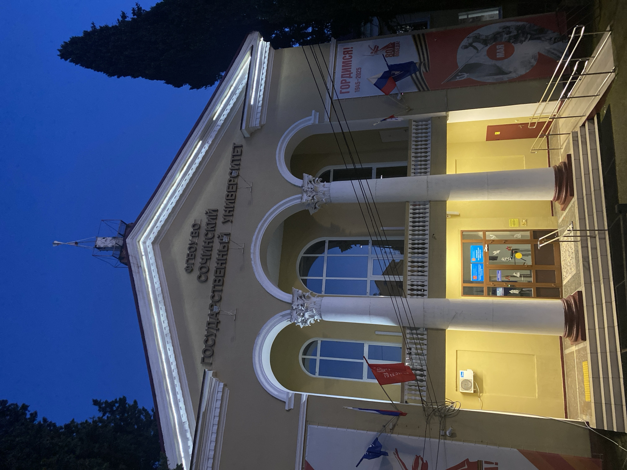 The image shows the facade of the Sochi State University building in Sochi, Russia, at dusk or night. The building has two stories, featuring architectural elements such as white columns, arches, and ornate details. The facade is illuminated with lights. Banners with Russian text and symbols related to Victory Day (May 9th) are displayed. The text on the building reads "ФГБОУ ВО Сочинский ГОСУДАРСТВЕННЫЙ УНИВЕРСИТЕТ" (FGBOU VO Sochi State University). Power lines are visible in the foreground. The sky is a dark blue. Trees are visible at the edge of the frame.
