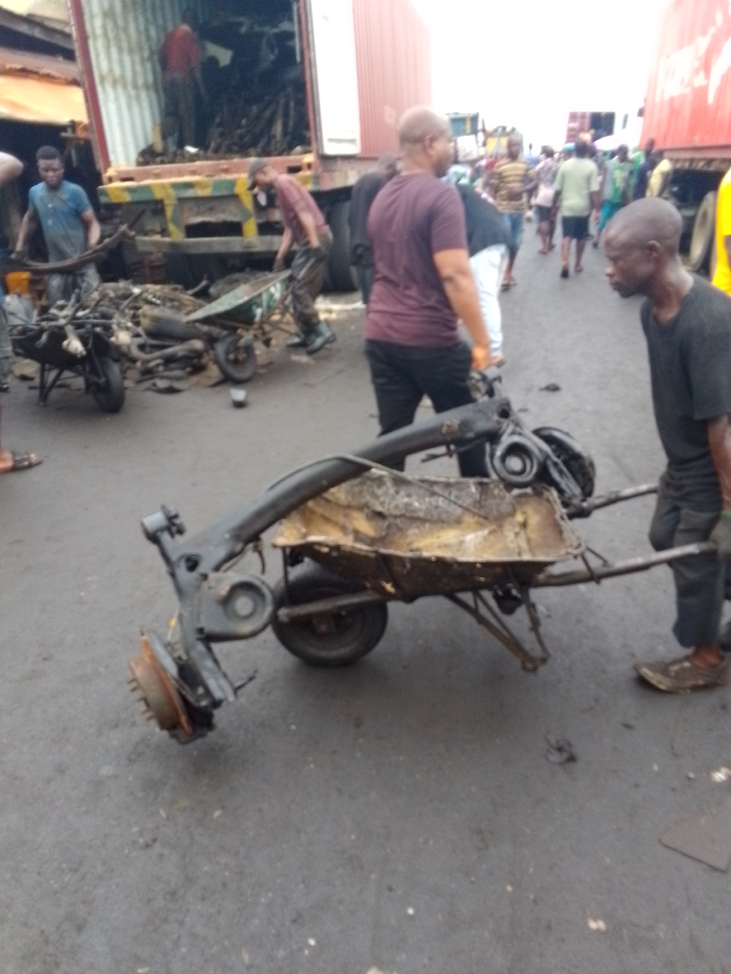 In Onitsha, Nigeria, a congested street scene shows several individuals engaged in manual labor near a large shipping container on a truck. The container appears to be filled with metal scrap. Some people are unloading the scrap, while others use wheelbarrows to transport it. In the foreground, a man hauls a heavily loaded wheelbarrow containing a car axle and other scrap metal. Pedestrians and additional trucks add to the chaotic environment. The street surface is asphalt and appears worn.