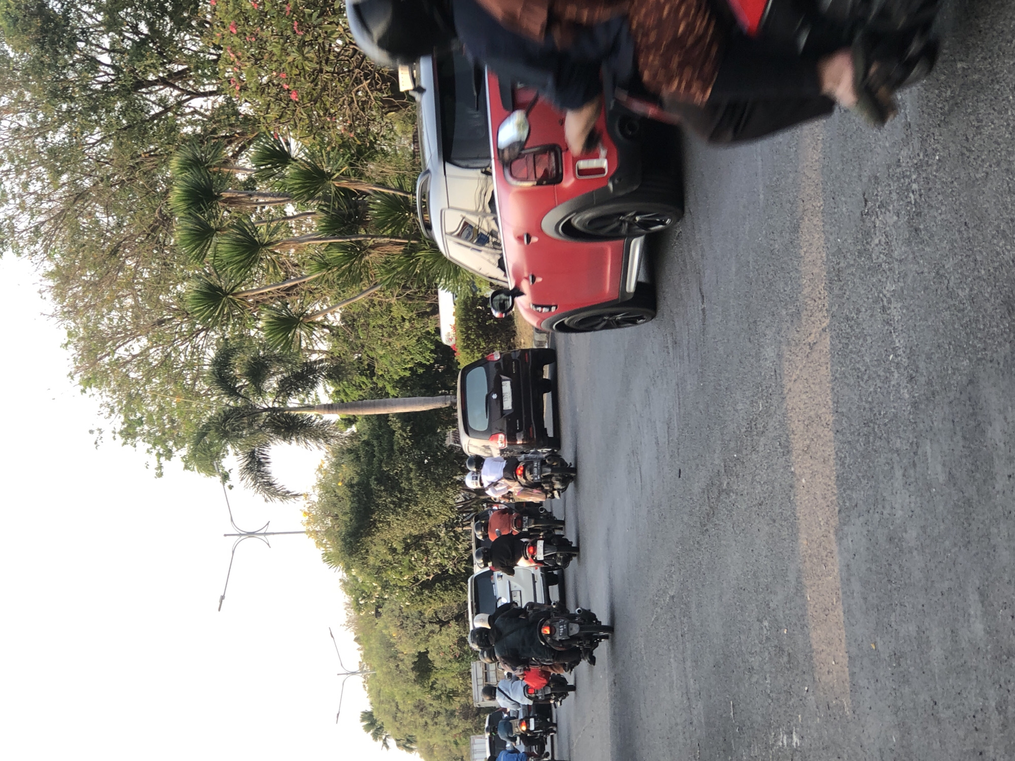 The image depicts a street scene in Ngaban, Indonesia, characterized by a moderate flow of traffic consisting primarily of motorcycles. In the foreground, a person on a red motorcycle is partially visible, wearing a dark helmet and a patterned garment. To the left, a red Mini Cooper is driving, its rear end facing the camera. Further down the road, several motorcycles are seen with riders, many wearing helmets. In the background, white vans and cars are also present, along with lush green trees and palm trees lining the street. Streetlights are visible in the distance. The asphalt road has a light brown stripe marking a lane. The overall atmosphere suggests a typical urban environment during daylight hours.