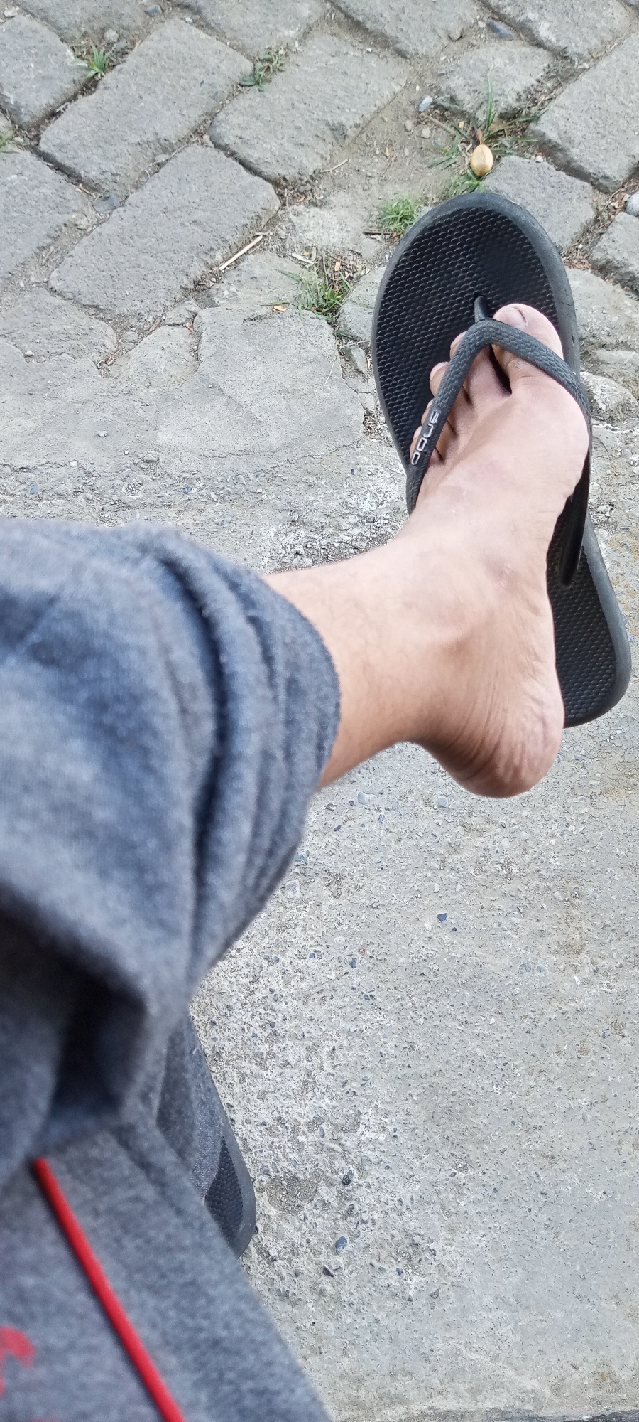 A person's bare foot wearing a black flip-flop is visible, positioned over gray paving stones. The foot is resting on a gray, textured surface. The person is wearing gray sweatpants with a red stripe. The environment appears to be an outdoor, paved area, possibly a sidewalk or pathway. There is some sparse greenery growing between the paving stones.
