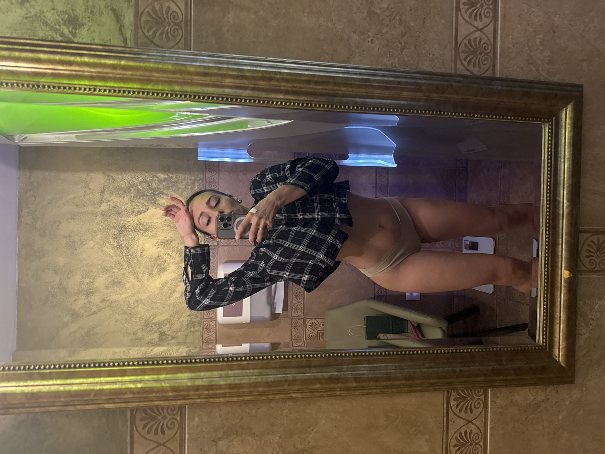 A young woman is captured in a mirror selfie, posing in what appears to be an indoor space such as a bathroom or changing room. She has light-colored hair and is wearing an open black and white plaid shirt, revealing light-colored underwear or briefs. Her left hand is raised, forming a peace sign, while her right hand holds a smartphone, likely an iPhone, which she is using to take the picture.
The mirror she is reflected in features a prominent, ornate gold frame with intricate beading details. The walls of the room are tiled in a warm, earthy brown or beige tone, adorned with decorative vertical strips featuring repeating Greek key-like or floral patterns.
In the reflection, a bright, luminous green object or light source is visible in the upper left, possibly indicating a tanning bed or a unique lighting fixture. Below the woman's arm, a white wall-mounted dispenser is present. On the floor behind her legs, a light-colored object resembling a bathroom scale can be seen, alongside a light-colored chair or bench with some items on it. The lighting is artificial, illuminating the interior space. The image is oriented sideways, giving the impression the woman is on her side. Her expression is neutral as she poses for the photograph. No discernible text is visible within the image. The location context provided is Saint Petersburg, Russia.