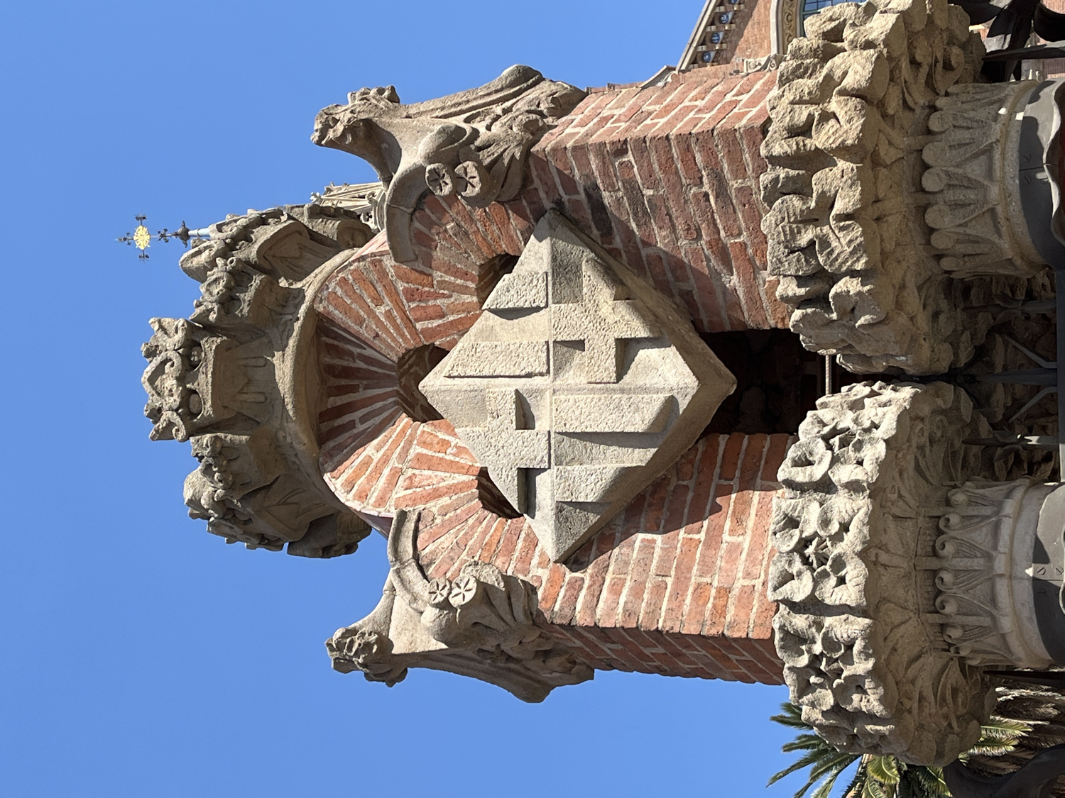 The image captures an upward view of intricate architectural details from a building's exterior in Barcelona, Spain, set against a clear, bright blue sky. The scene is illuminated by strong sunlight, casting crisp shadows that emphasize the three-dimensional nature of the construction.
At the core of the composition is a prominent, light-colored stone element, shaped like a rhomboid or square placed on its corner. Its visible face is carved with a complex, relief pattern resembling a stylized cross or geometric steps. This stone piece is integrated into a structure primarily built from reddish-brown bricks, laid in decorative patterns that include radiating, fan-like arches and straight courses.
Surrounding the brickwork and central stone are highly ornate, carved stone elements. These appear to be decorative capitals, corbels, or structural supports. They feature rich, sculptural details, including what look like grotesque or gargoyle-like figures with intricate facial features, along with elaborate floral motifs, abstract patterns, and organic forms. One visible cylindrical section is densely carved with repeating floral or rosette-like designs.
In the upper left quadrant, a small, gilded finial or weather vane, resembling a cross with four arms, is visible against the sky, indicating the very top of a spire or decorative element. The lighting suggests it is a clear, sunny day, likely mid-morning or early afternoon. The fronds of a palm tree can be seen in the lower right, subtly indicating a warm, possibly Mediterranean, climate. There is no visible text in the image. The architectural style is highly decorative, characteristic of Modernisme, a prominent movement in Barcelona.